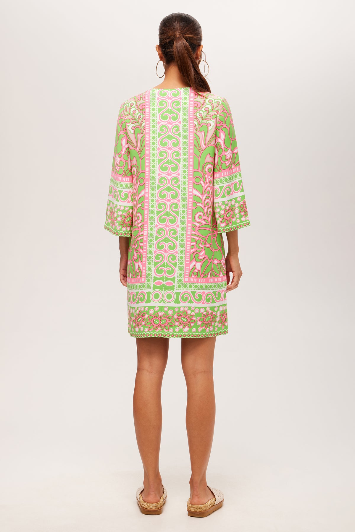 A woman with long brown hair tied back wears hoop earrings, flat sandals, and the Botanical 3/4-Sleeve Zip Dress - Petra Floral featuring green, pink, and white geometric and floral patterns. She stands facing away.