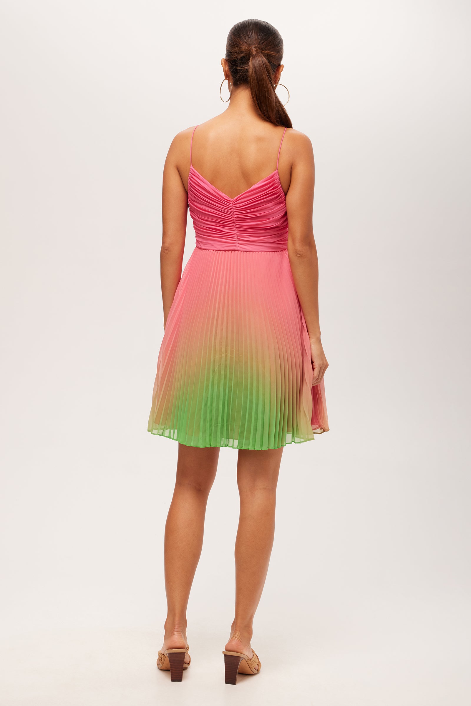 A woman with long brown hair in a ponytail wears the Natalie 2 Sleeveless Pleated Ombre Dress, featuring thin straps and a pink-to-green gradient. She stands with her back to the camera against a plain background.