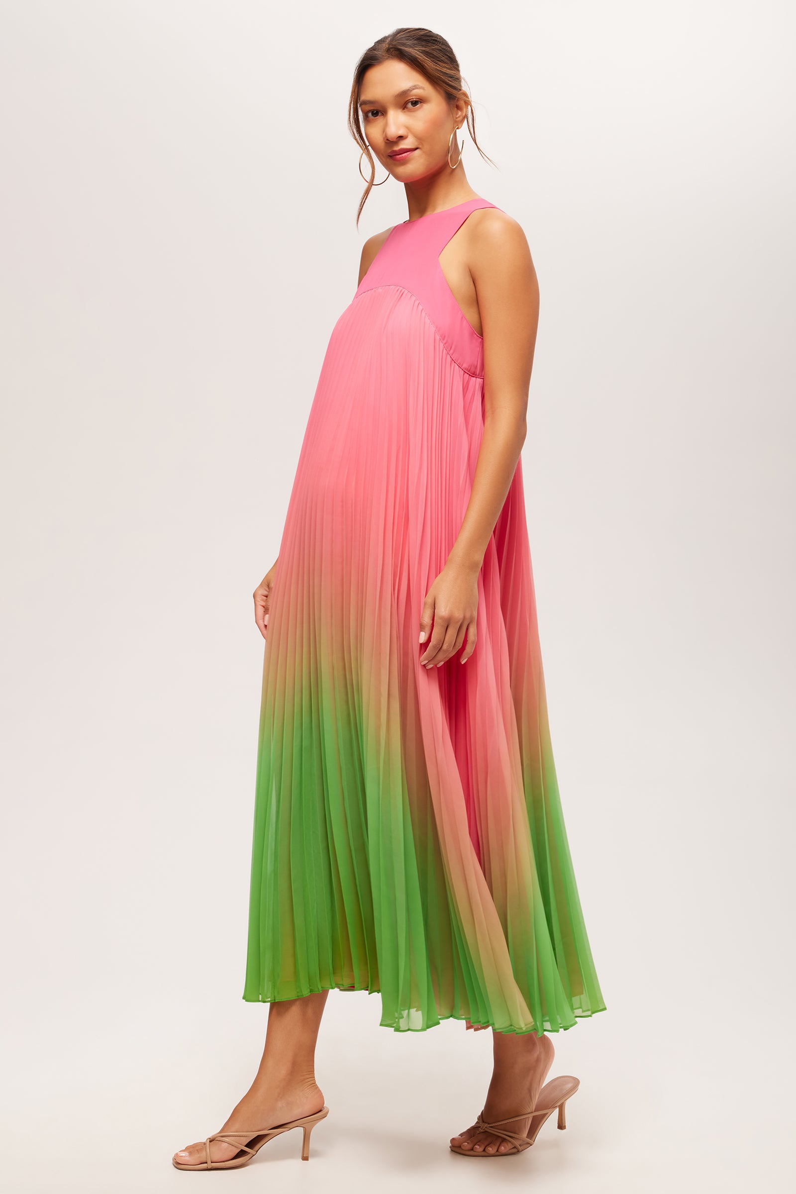 A woman poses against a plain light background in the Jalina 2 Sleeveless Pleated A-Line Midi Dress, featuring a pink bodice that fades into green at the hem. She completes the look with nude heeled sandals and her hair tied back.