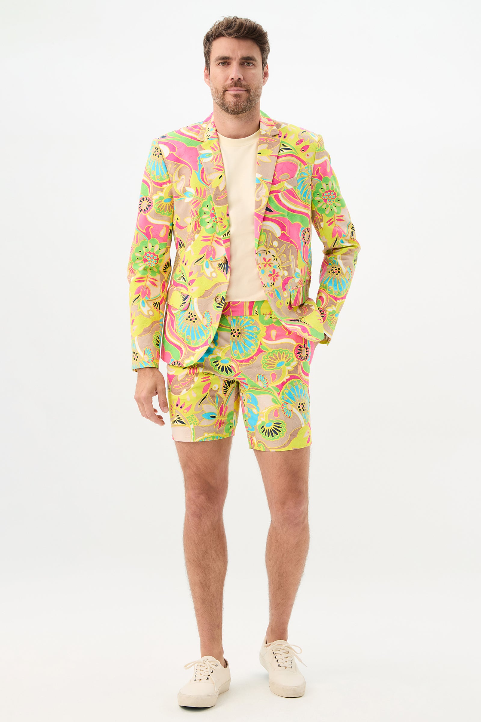 A man stands against a white background wearing the Thurston Blazer - Venus, styled with matching shorts, a light beige T-shirt, and white sneakers. The suit showcases bold pink, yellow, blue, and green abstract patterns.