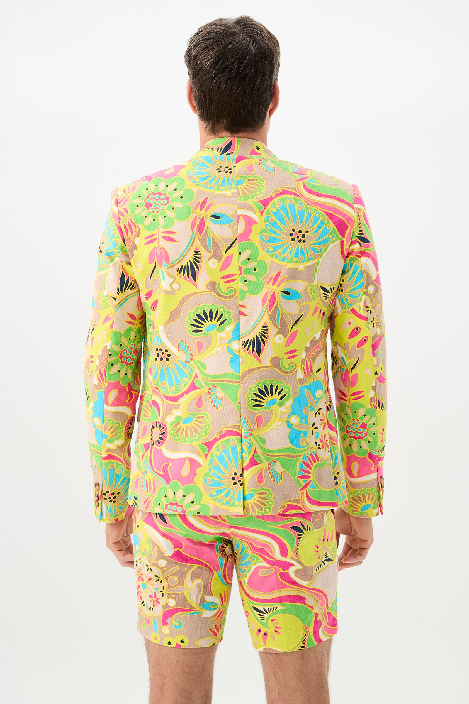 A man stands facing away, wearing the Thurston Blazer - Venus: a colorful suit jacket and matching shorts featuring a bold neon pink, green, yellow, and blue floral and abstract pattern on a light background.
