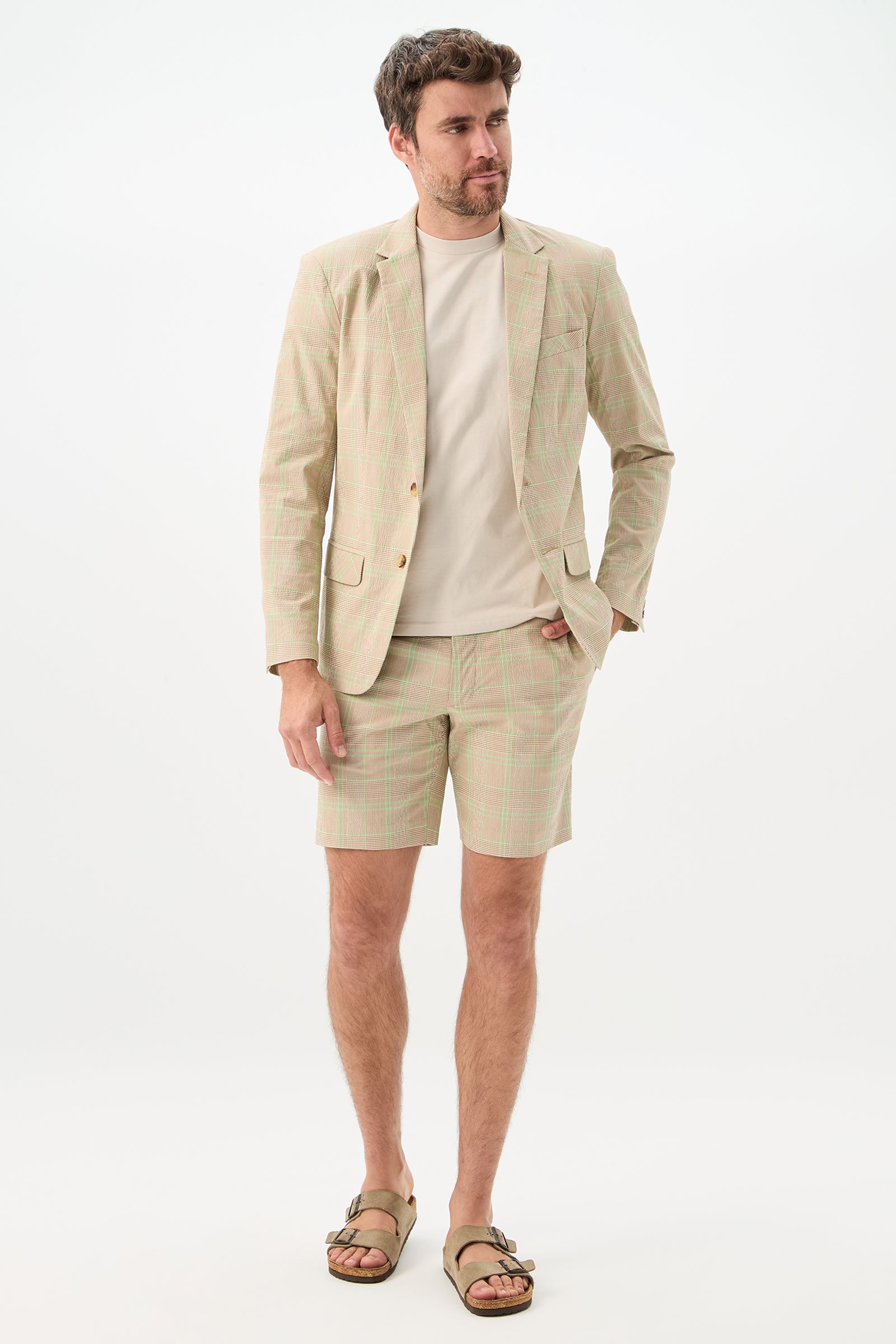 A man wears the Louie Blazer - Zianieri Plaid with matching shorts, a light beige t-shirt, and tan sandals, standing against a plain white background with a relaxed expression and one hand in his pocket.