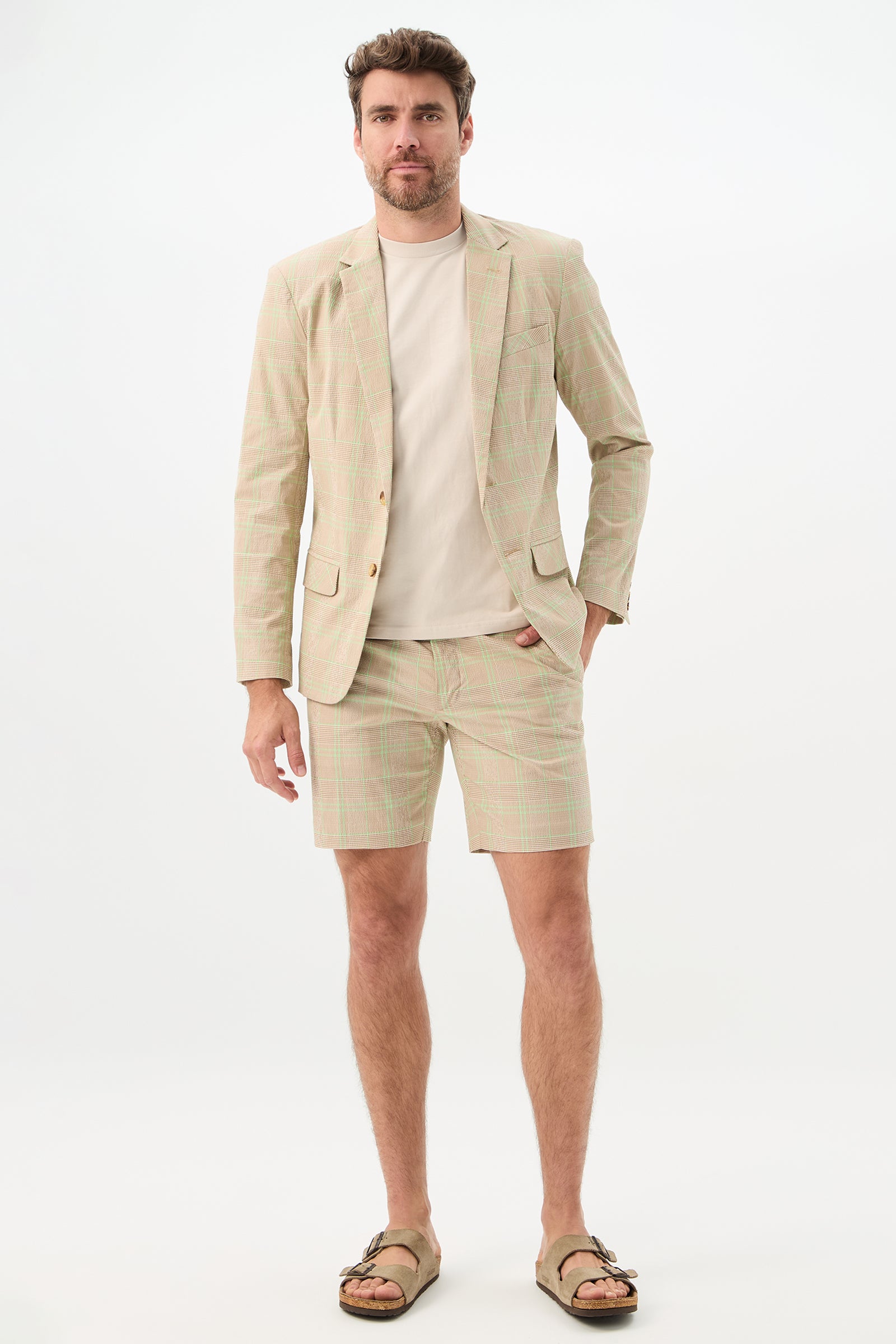 A man with a neutral expression stands against a white background wearing the Travis Short - Zianieri Plaid, paired with a matching blazer, beige t-shirt, and tan sandals, one hand in his pocket.