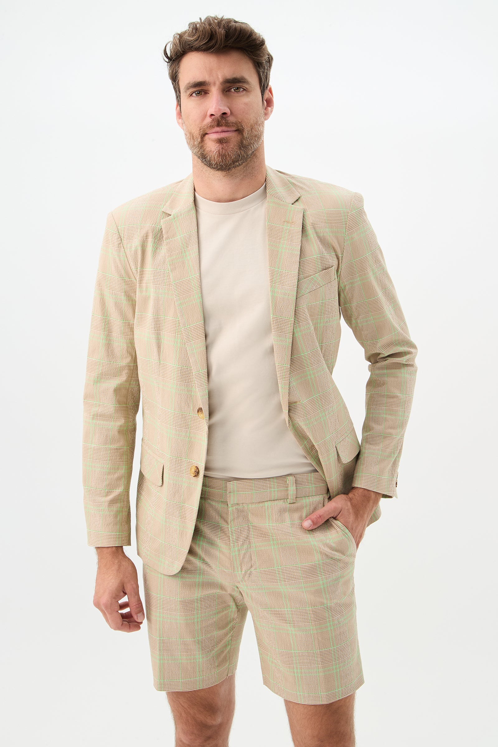 A man with brown hair and a beard wears the Louie Blazer - Zianieri Plaid in Beach, paired with matching shorts and a cream t-shirt. He stands against a white background, one hand in his pocket, smiling slightly.