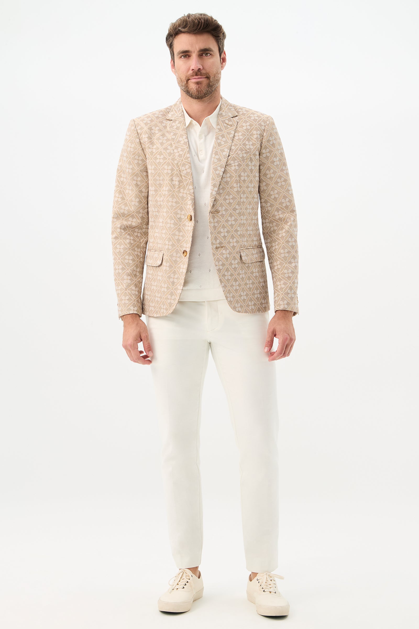 A man with short brown hair and a beard stands against a white background, wearing the Thurston Blazer - Metallic Daisy Jacquard over a white shirt, paired with white pants and sneakers. He looks neutral with hands relaxed by his sides.