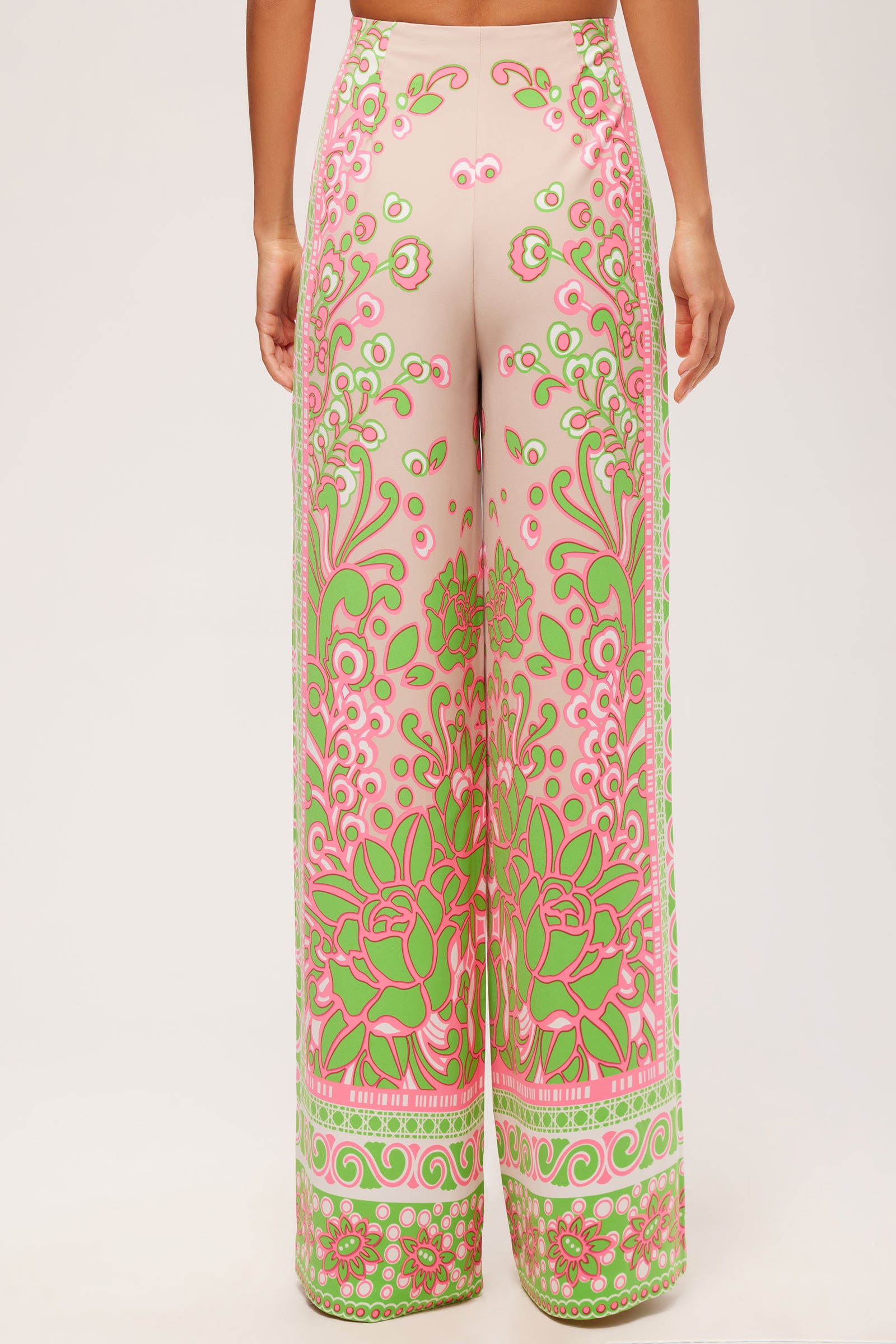 A person stands with their back to the camera, wearing the Federer Wide-Leg Pant - Petra Floral: high-waisted, floor-length pants with a pink base and detailed green and pink floral patterns at the waistband and legs.