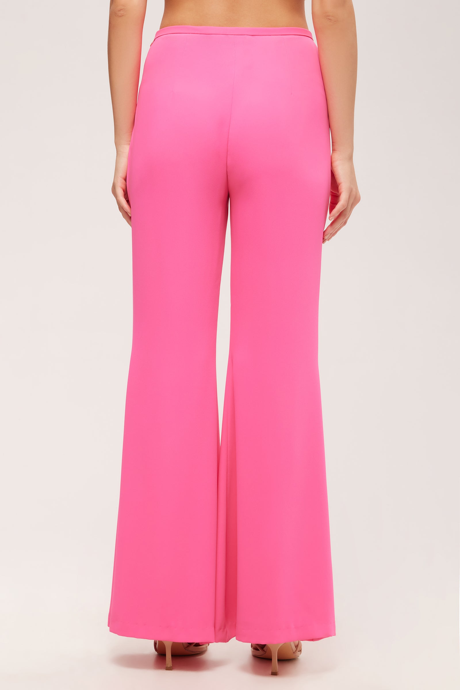 A woman is seen from behind wearing the Drop Shot Flared Pant - Double Georgette, a high-waisted, bright pink, wide-leg pant, standing against a plain light background.