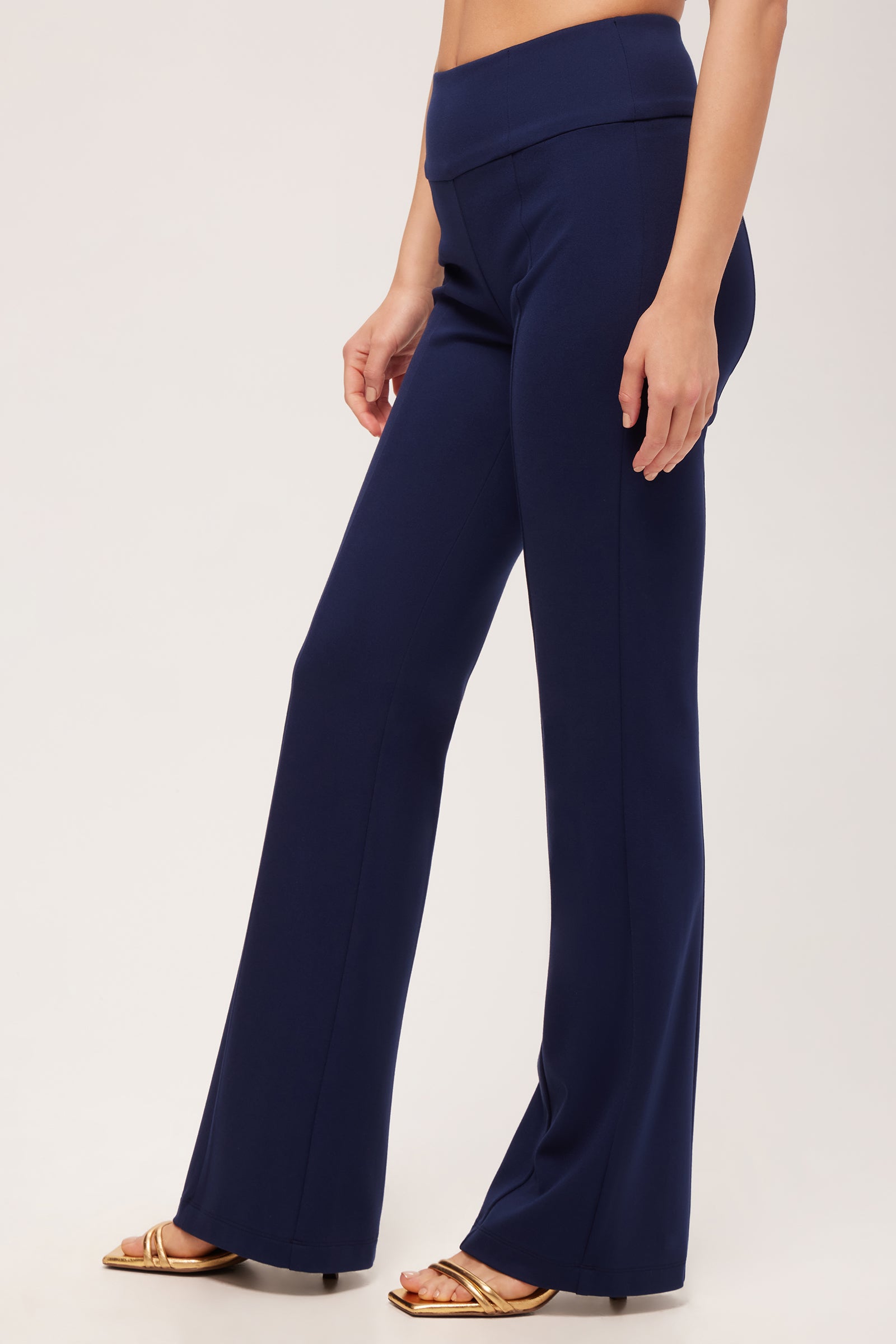 A person is shown from the waist down, standing against a plain light background, wearing Becker High-Waist Flared Pant - Cozy Ponte in navy blue and gold sandals.