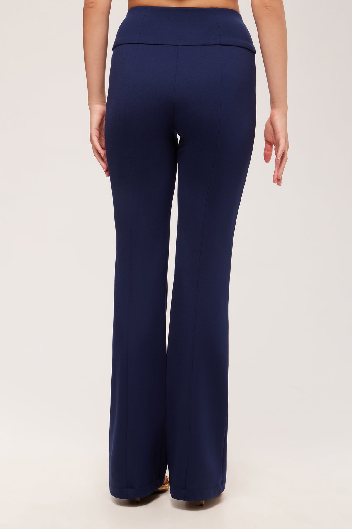 A person faces away from the camera, wearing the Becker High-Waist Flared Pant - Cozy Ponte in navy blue with beige shoes against a plain, light-colored background.