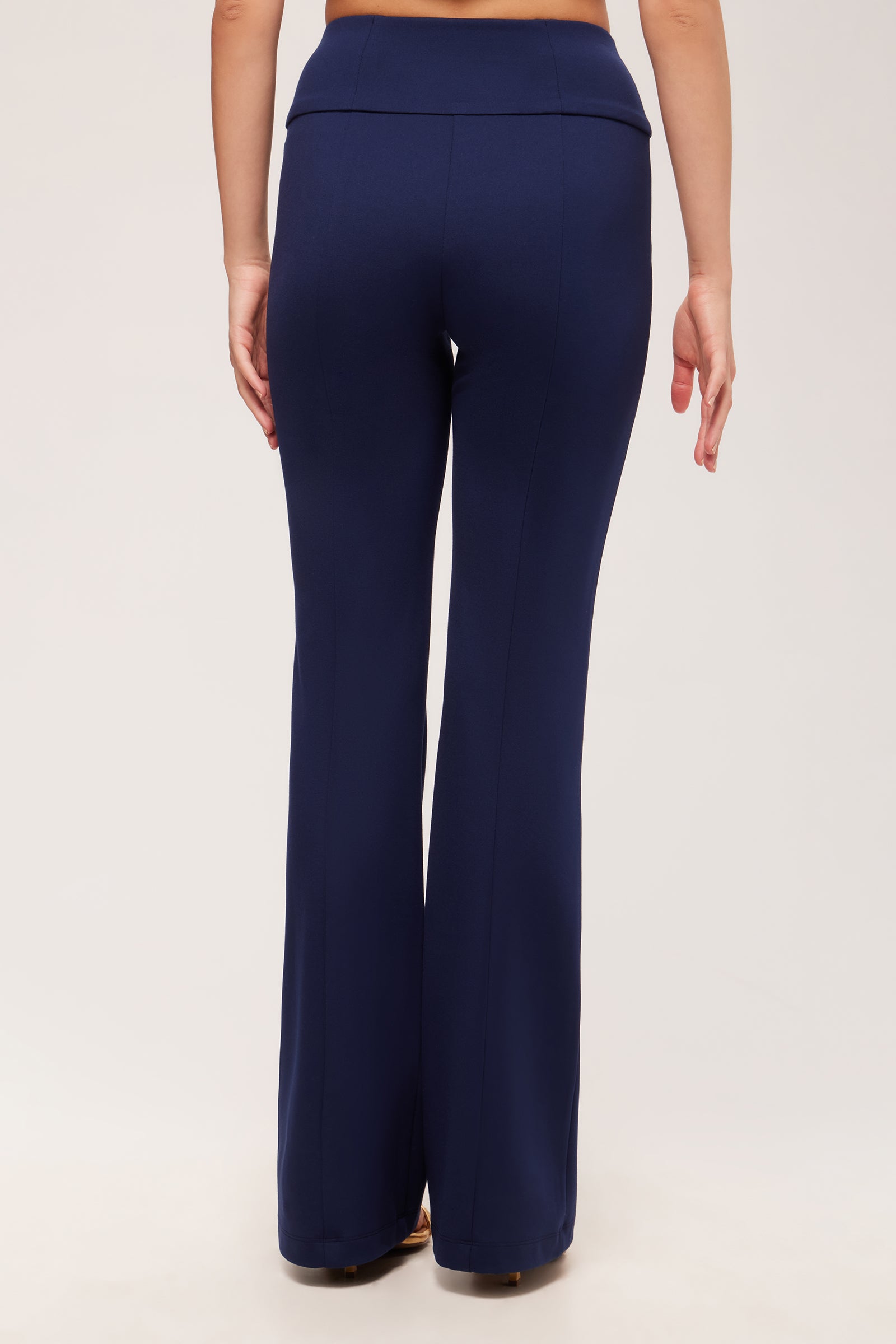 A person faces away from the camera, wearing the Becker High-Waist Flared Pant - Cozy Ponte in navy blue with beige shoes against a plain, light-colored background.
