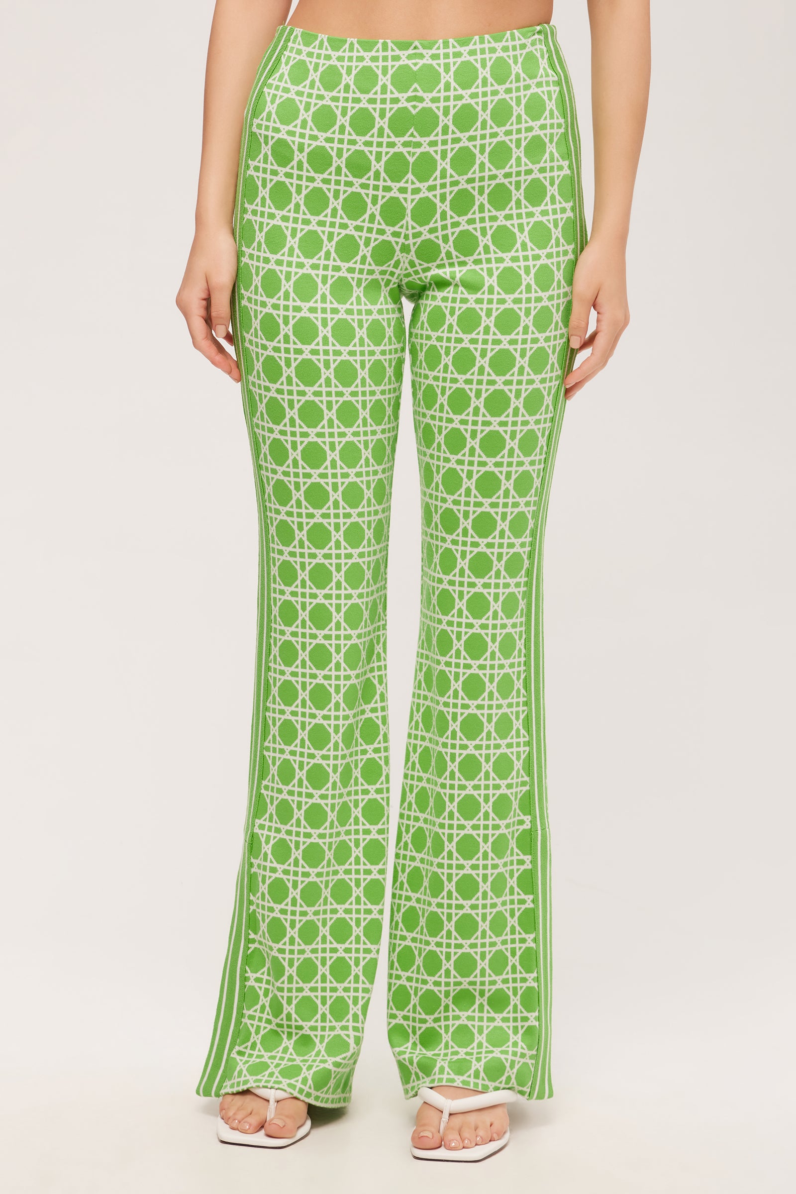 A person wearing the Tennis Bootcut Pant in Billie Jean stands against a plain light background, paired with white open-toe heels. The green high-waisted pants feature a white geometric pattern, and the image is cropped at the midriff.