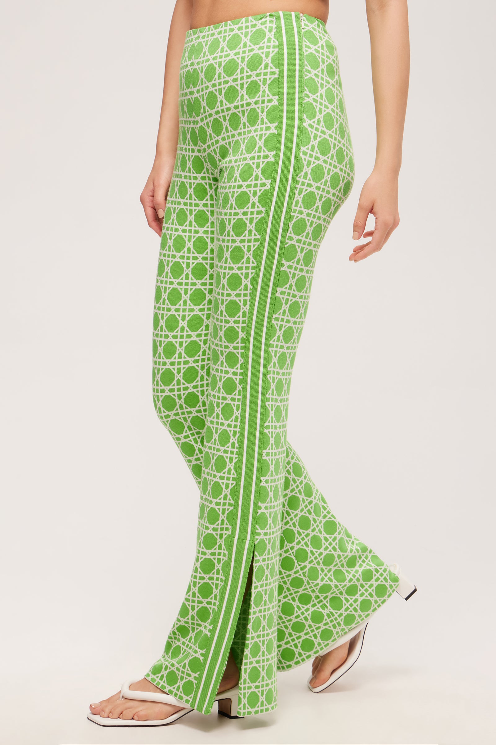 A person wears the Tennis Bootcut Pant - Billie Jean, featuring green and white patterns with a side stripe, paired with white open-toe sandals, against a plain light background.