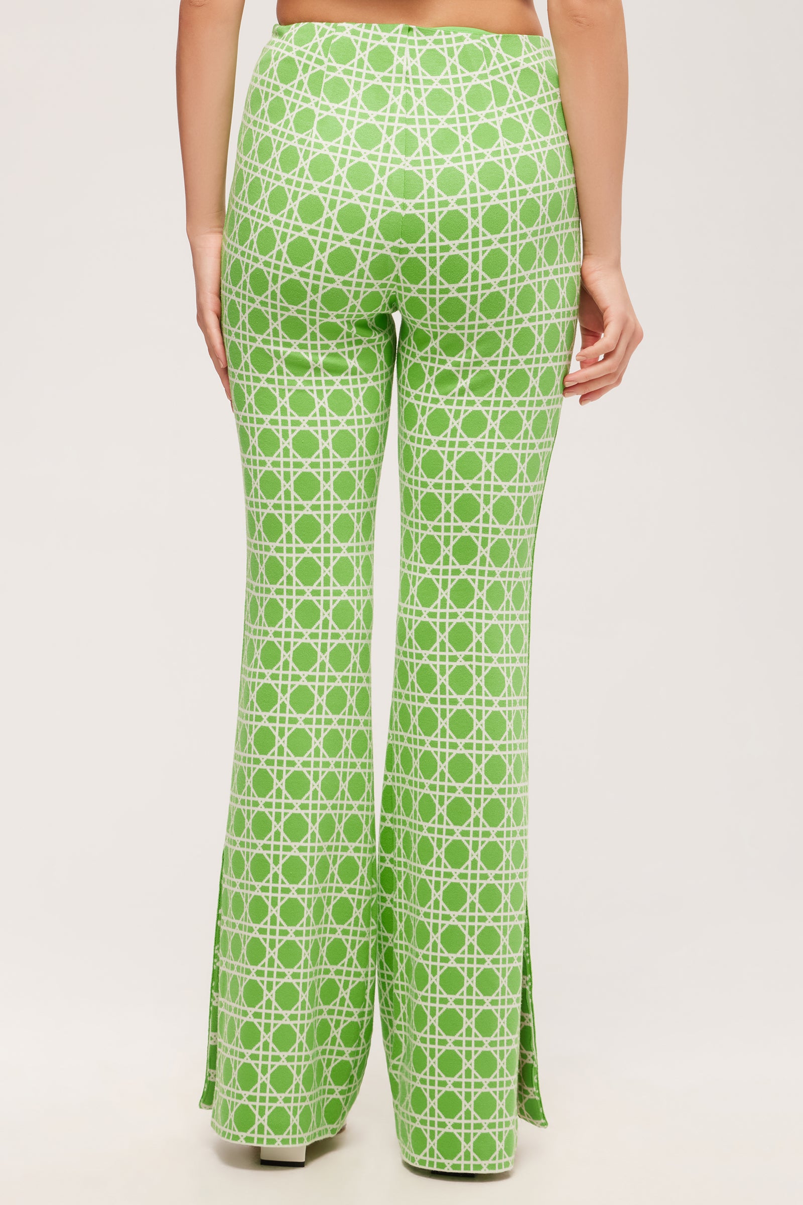 A person is shown from the waist down, wearing the Tennis Bootcut Pant - Billie Jean: high-waisted, green bell-bottoms with a white geometric pattern and side slits. The background is plain and light-colored.
