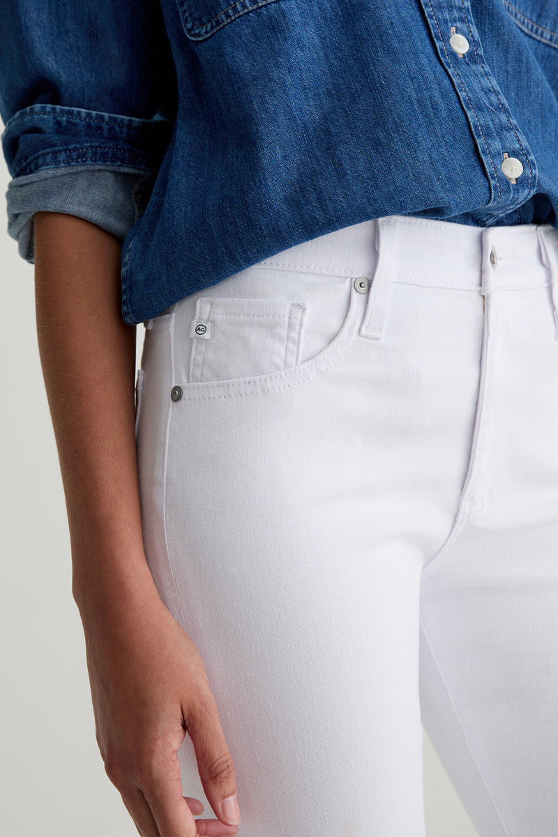 A person shown from mid-torso to upper thigh wears a blue denim shirt with rolled-up sleeves and AG Farrah Mid-Rise Skinny Ankle Jean in white, their left hand relaxed by their side.