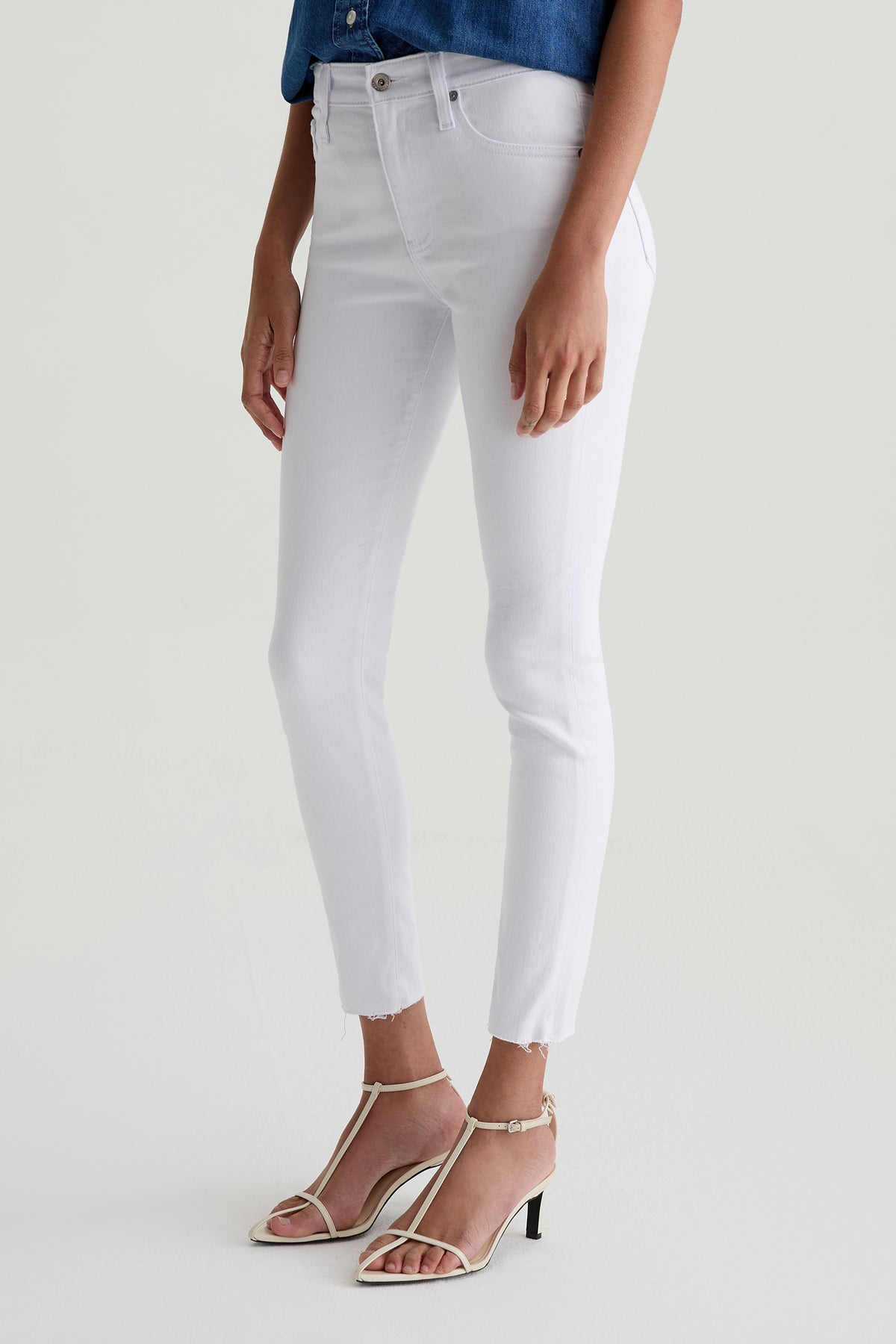 A person wearing AG Farrah Mid-Rise Skinny Ankle Jean in white, a blue denim shirt, and white heeled sandals stands against a plain light background. The image displays their body from shoulders down.