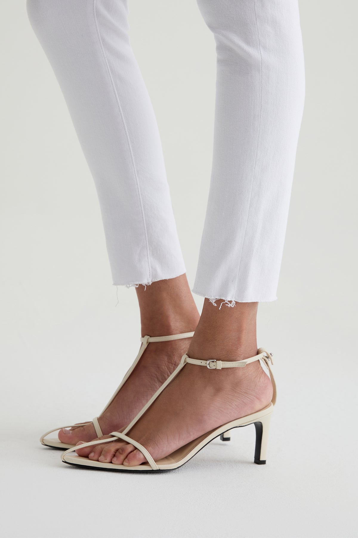A person wearing the AG Farrah Mid-Rise Skinny Ankle Jean in white with beige, strappy heeled sandals featuring a thin ankle strap, standing against a light background.
