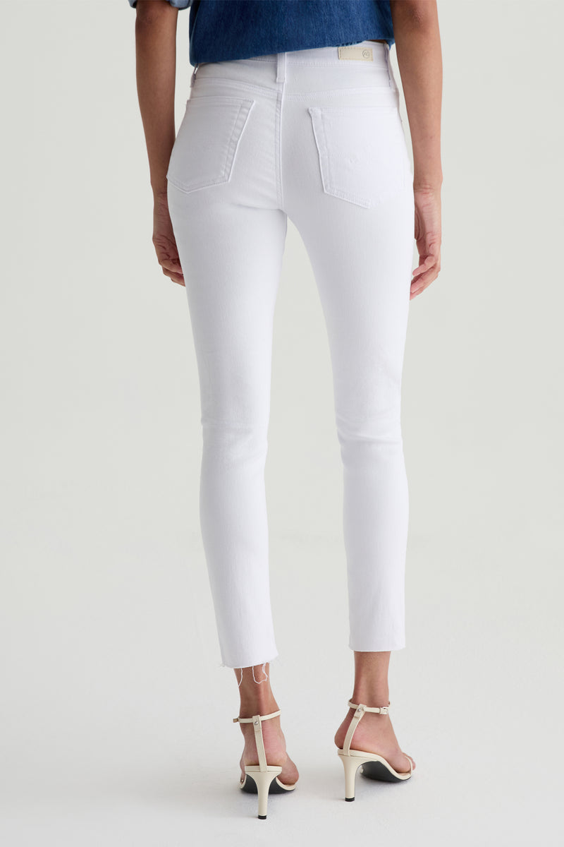 A person wearing AG Farrah Mid-Rise Skinny Ankle Jean in white with back pockets and a raw hem, paired with white heeled sandals and a blue short-sleeve top, is standing facing away from the camera.