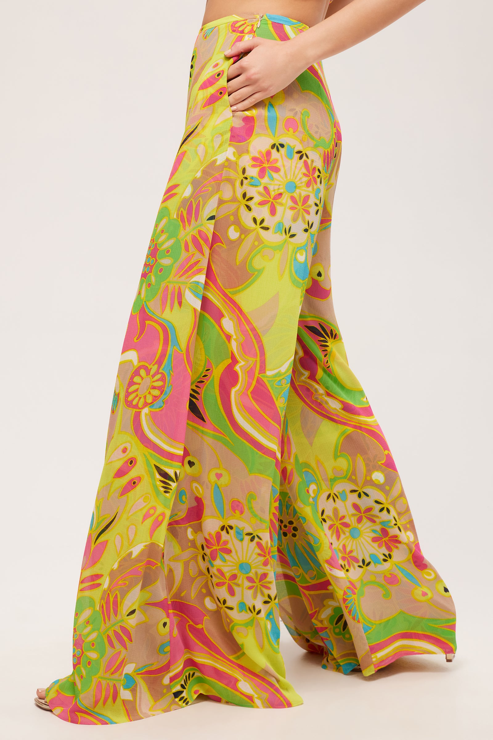 A person wears the Jack Wide-Leg Pant - Venus, featuring a high-waisted, wide-leg fit and a colorful retro floral pattern in yellow, green, pink, and orange. Hand on hip; plain white background.