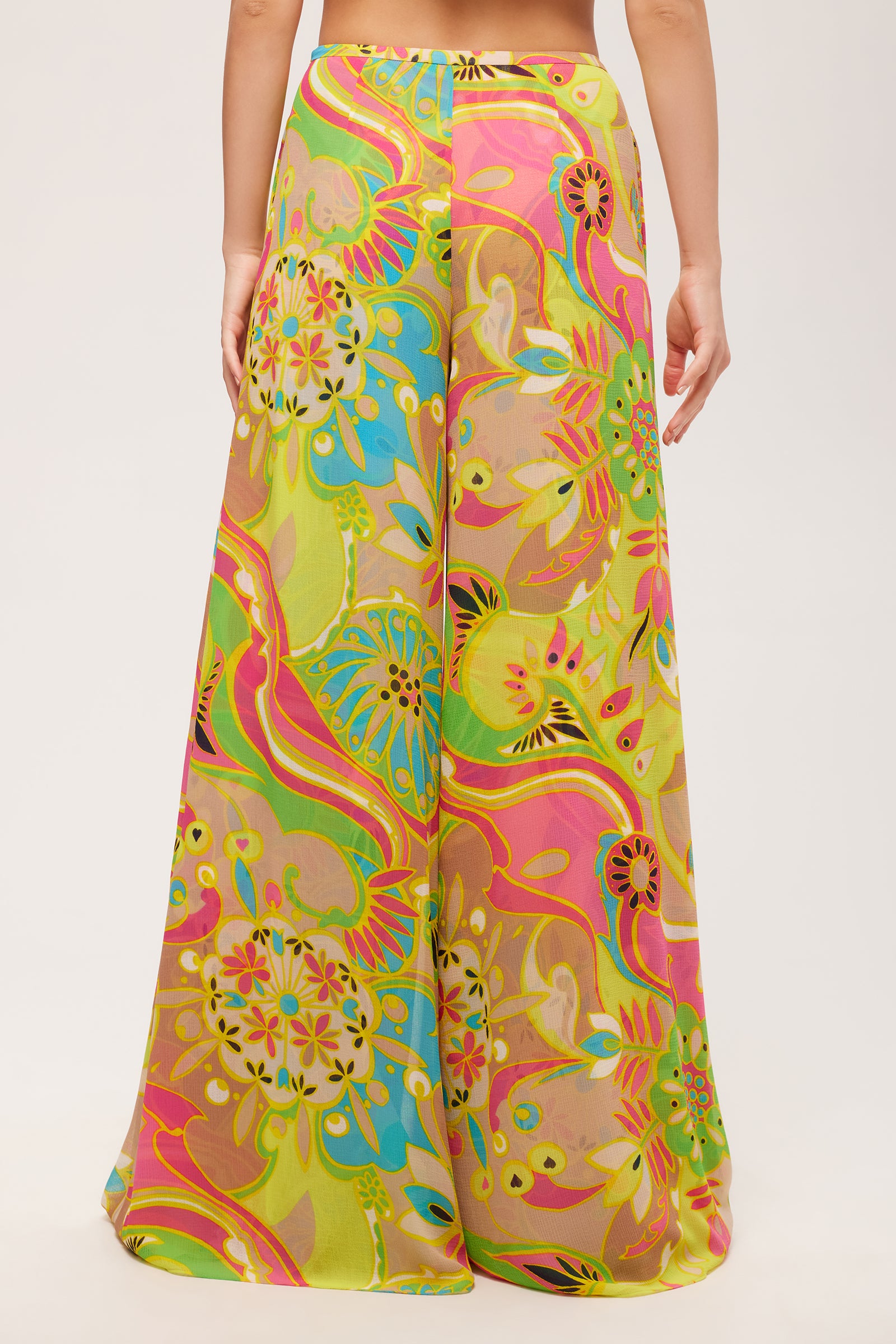 A person stands facing away, wearing the Jack Wide-Leg Pant - Venus, featuring a high-waisted fit and a vibrant multicolored floral and abstract print in yellow, pink, green, and blue against a plain white background.