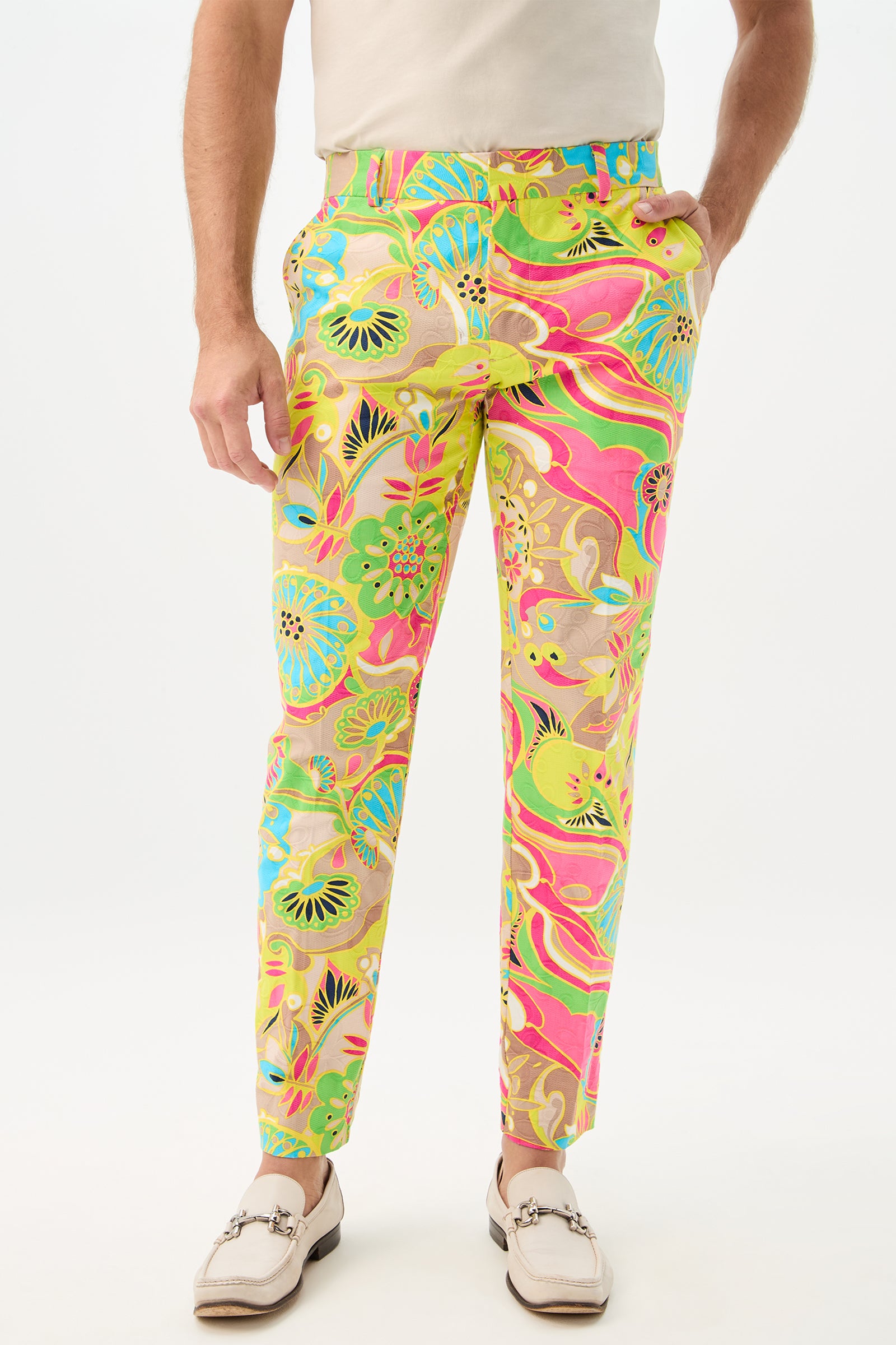 Someone is wearing the Clyde Slim Trouser in Venus, featuring colorful green, pink, blue, and yellow patterns, paired with a plain white shirt and light-colored loafers, standing against a white background.