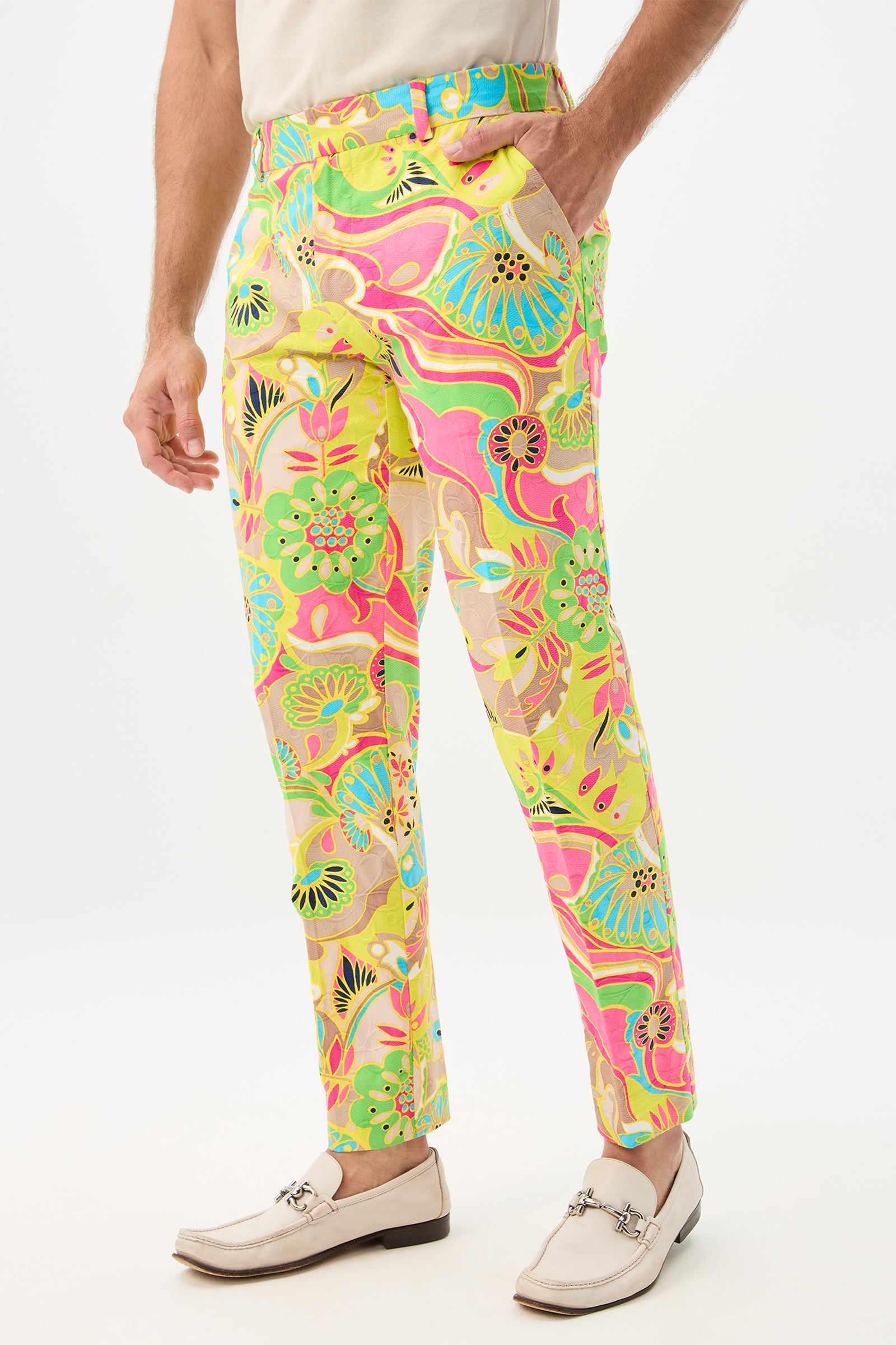 A person wears the Clyde Slim Trouser - Venus, featuring a bold floral and abstract pattern in pink, yellow, blue, and green, paired with a white shirt and light loafers. One hand rests casually in their pocket.