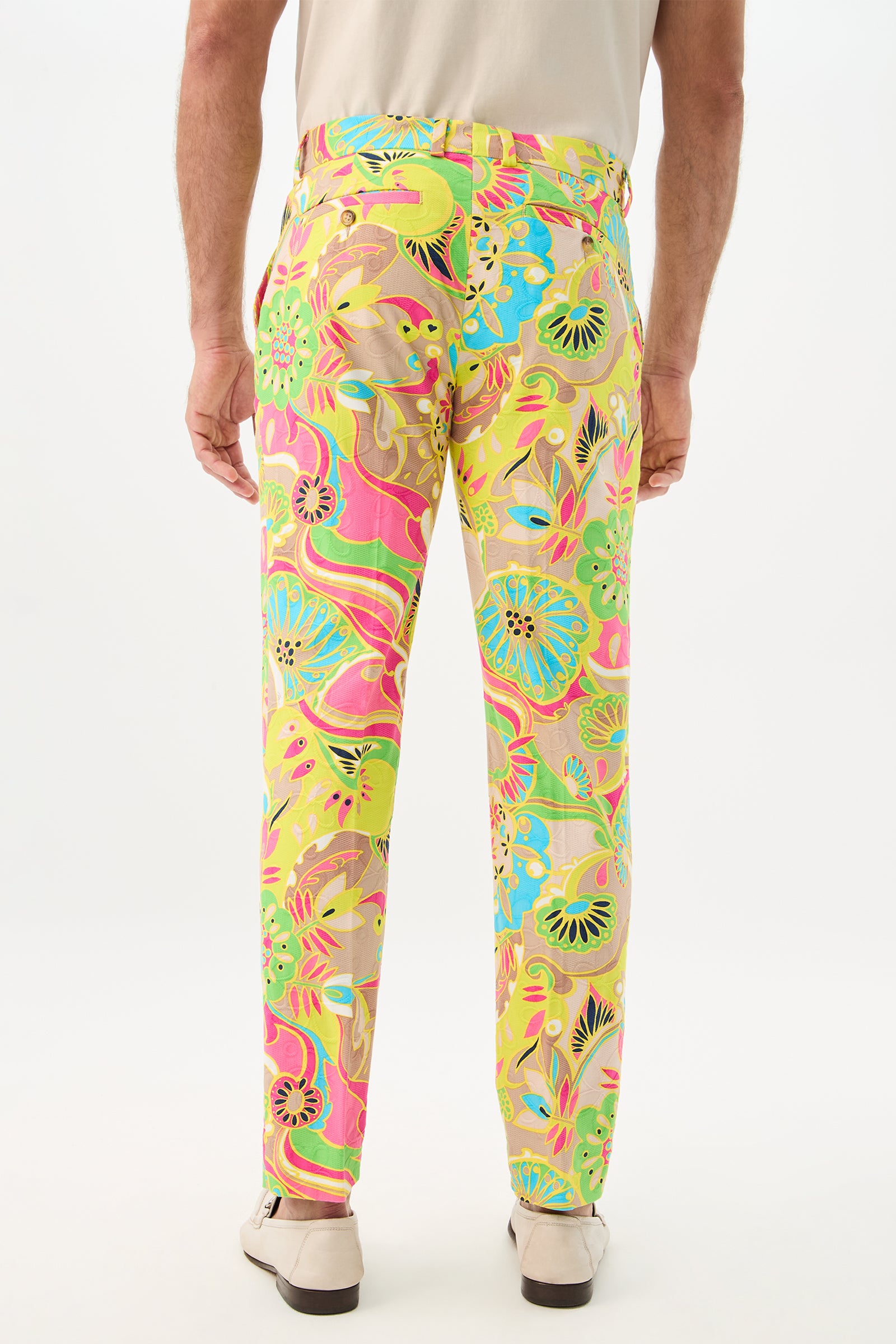 A person is seen from behind wearing beige shoes and Clyde Slim Trouser - Venus, featuring a bright, swirling floral pattern in pink, yellow, green, and blue against a plain white background.