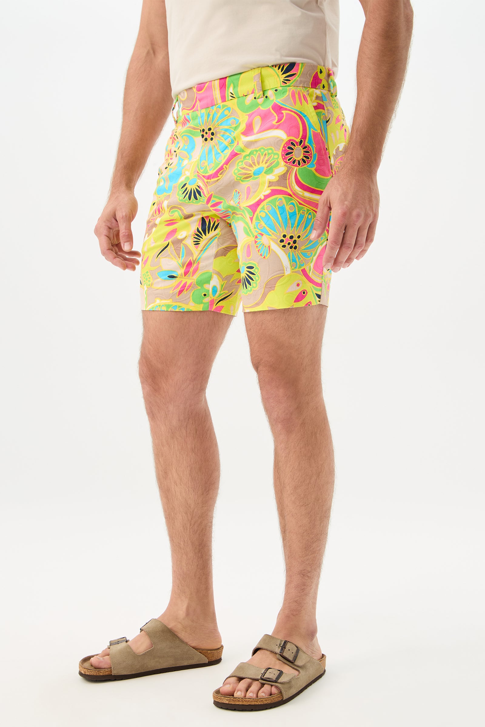 A person models the Lawrence Short - Venus, featuring bright, colorful florals, with a beige shirt and tan sandals against a white background. Only the lower body is shown.