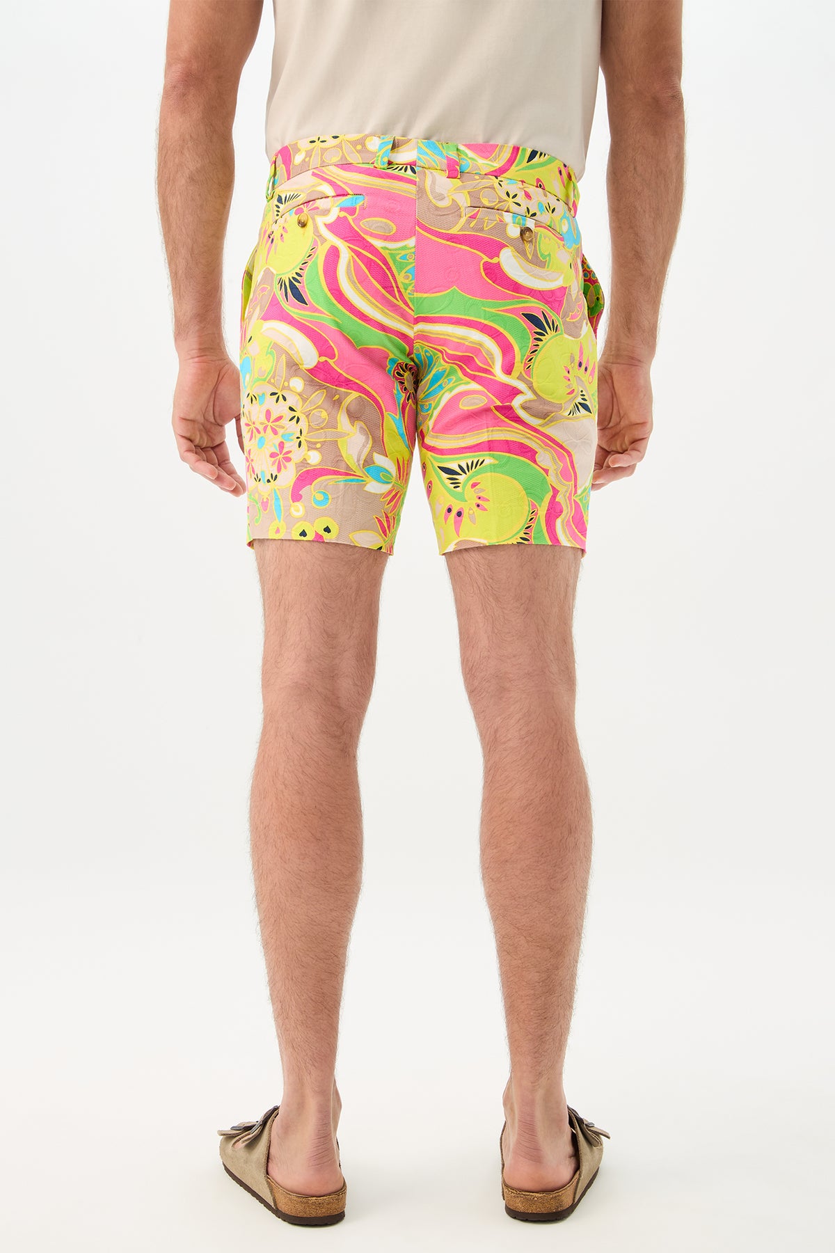 A man stands facing away on a white background, wearing the Lawrence Short - Venus, featuring colorful abstract patterns in pink, green, and yellow, paired with a beige short-sleeve shirt and brown slip-on shoes.
