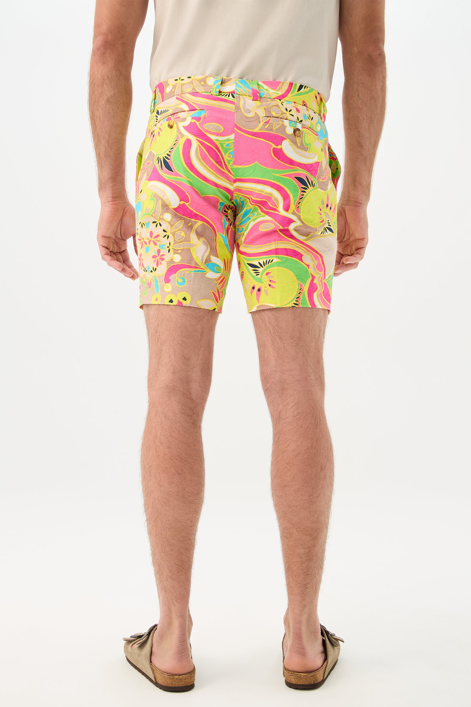 A man stands facing away on a white background, wearing the Lawrence Short - Venus, featuring colorful abstract patterns in pink, green, and yellow, paired with a beige short-sleeve shirt and brown slip-on shoes.