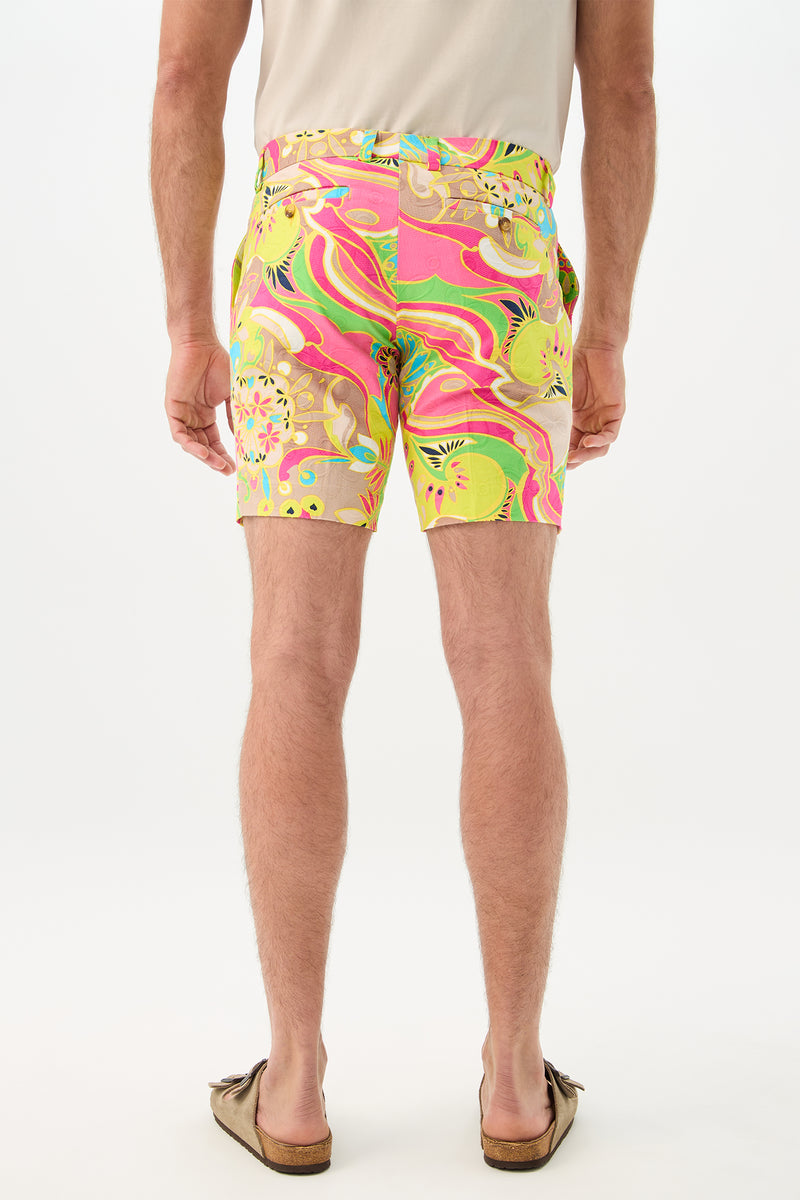 A man stands facing away on a white background, wearing the Lawrence Short - Venus, featuring colorful abstract patterns in pink, green, and yellow, paired with a beige short-sleeve shirt and brown slip-on shoes.