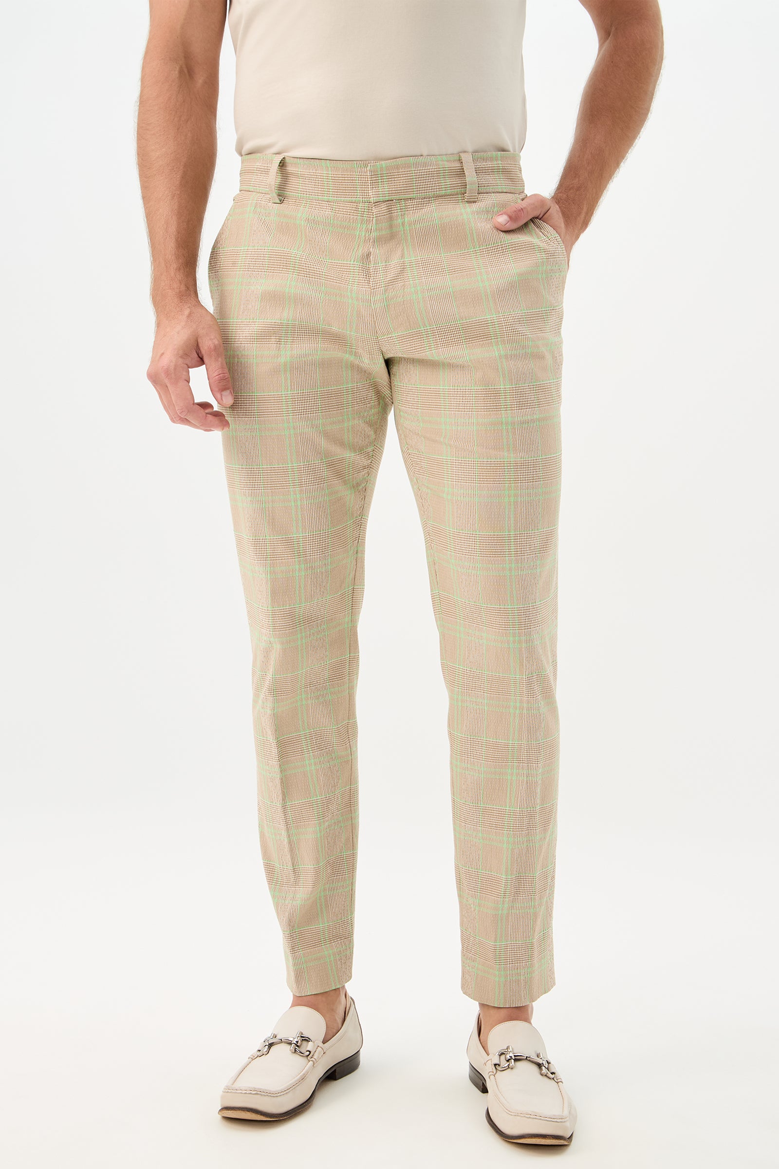 Someone stands against a plain white background, wearing the Clyde Slim Trouser - Zianieri Plaid in Beach, paired with a cream shirt and white loafers, one hand in their pocket.