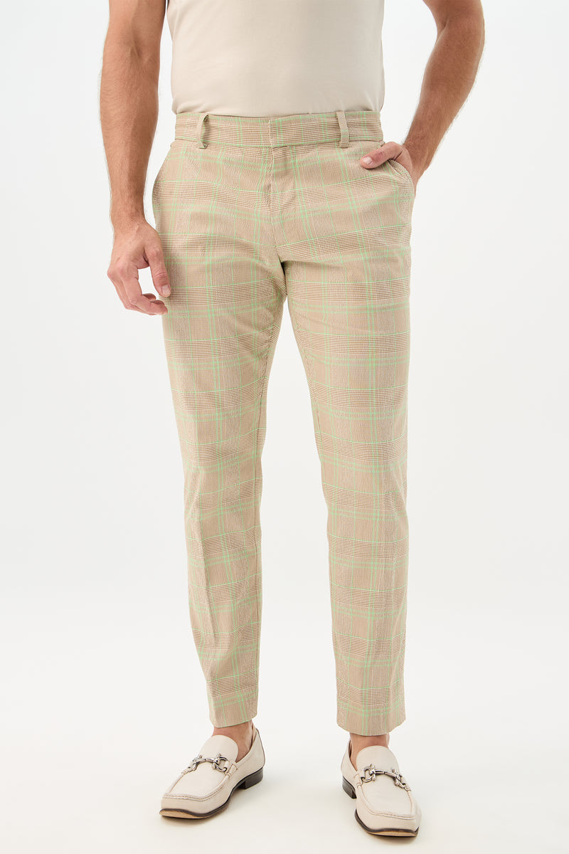 Someone stands against a plain white background, wearing the Clyde Slim Trouser - Zianieri Plaid in Beach, paired with a cream shirt and white loafers, one hand in their pocket.