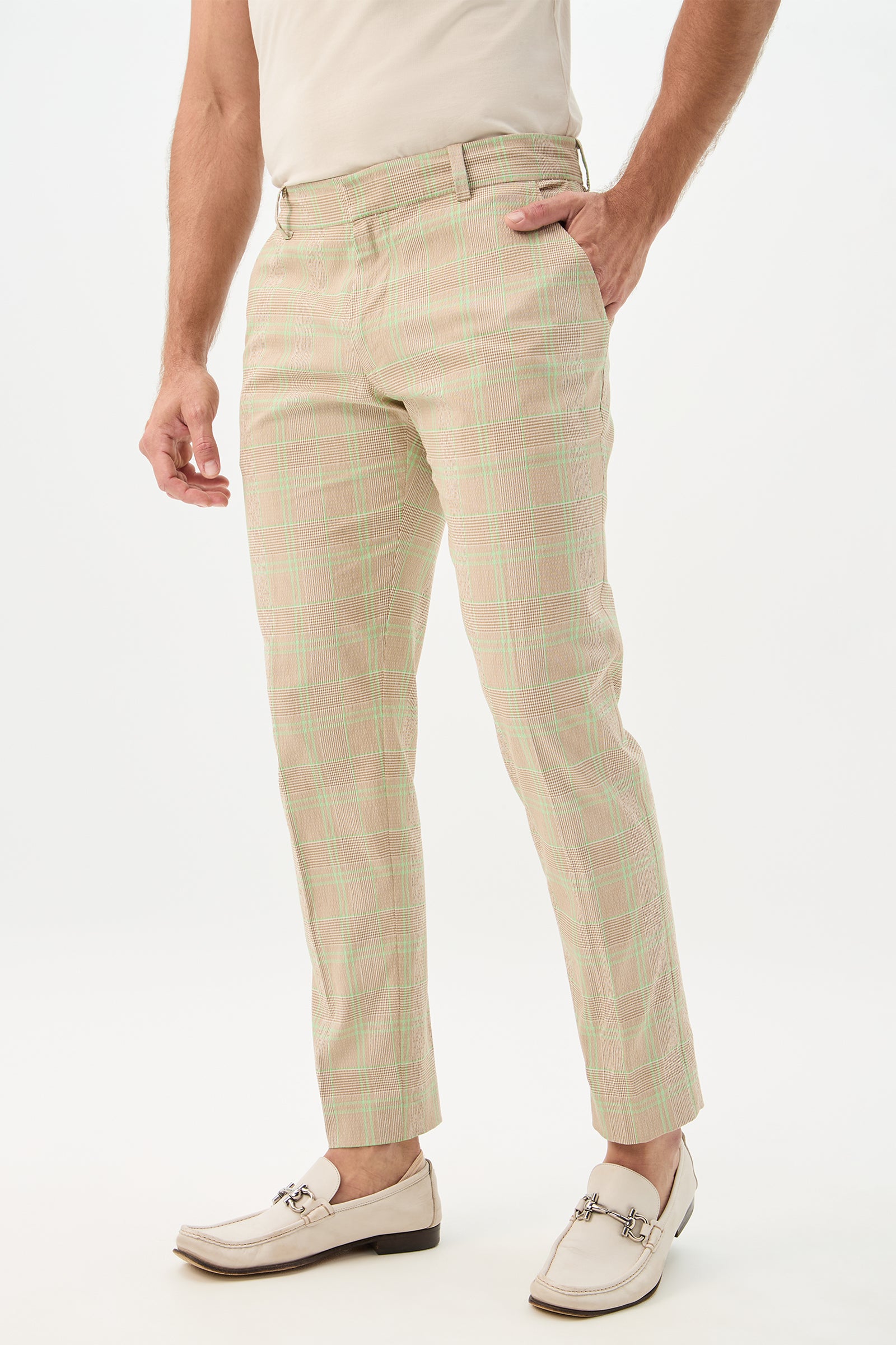 A man wearing the Clyde Slim Trouser - Zianieri Plaid, featuring a light pink and green checked pattern, paired with a beige shirt, cream belt, and white loafers with metal detail, stands against a plain white background.