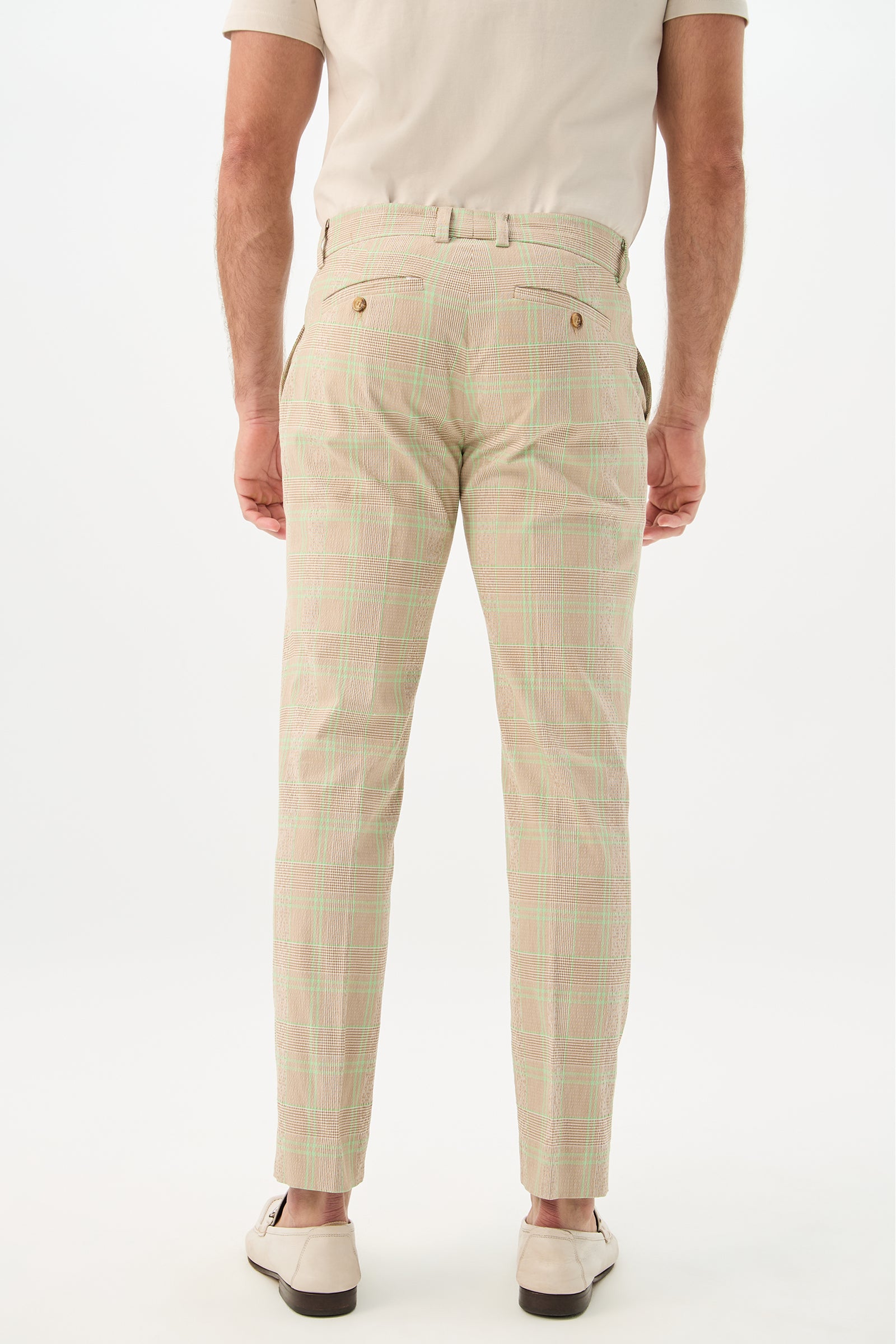 A man stands facing away, wearing Clyde Slim Trouser - Zianieri Plaid with green and pink lines, a beige short-sleeve shirt, brown belt, and white loafers against a plain white background.