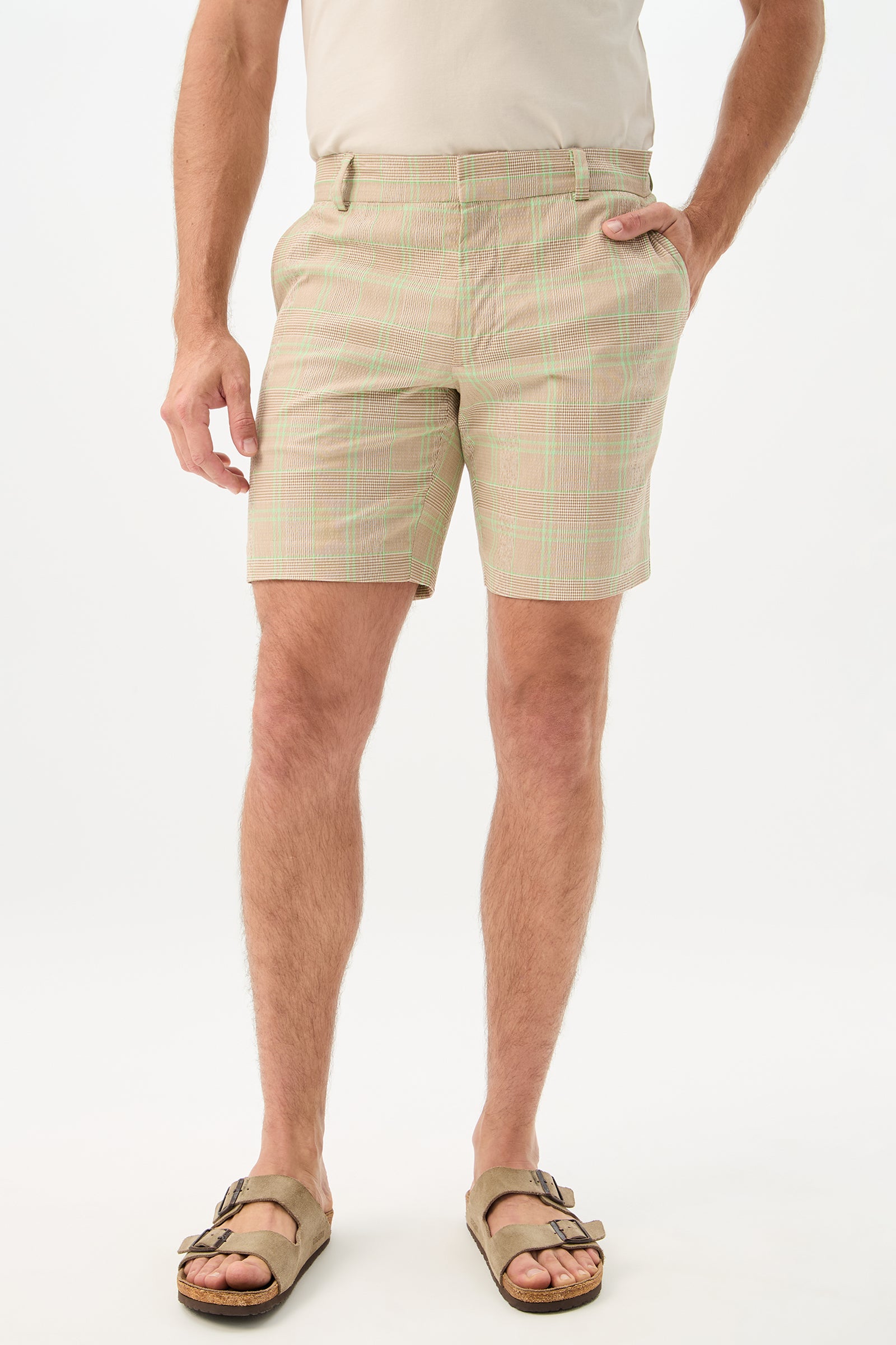 A man stands against a plain white background with one hand in his pocket, wearing the Travis Short - Zianieri Plaid in Beach color, paired with a beige t-shirt and tan sandals.