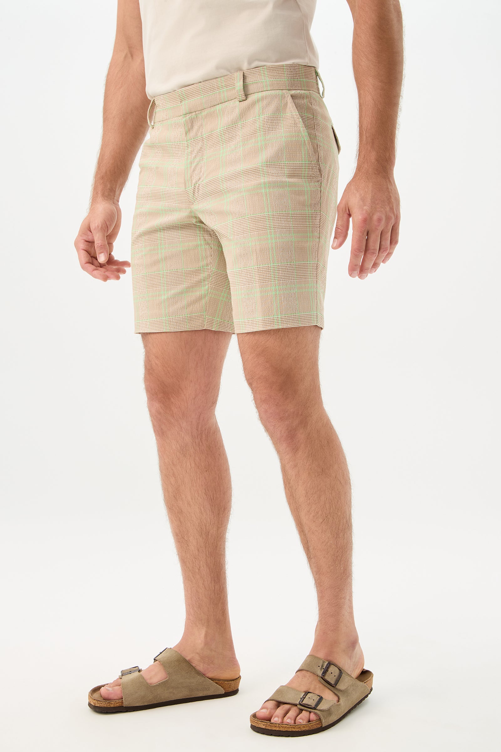 A person stands against a plain white background wearing the Travis Short - Zianieri Plaid in light beige and green, a tucked-in white shirt, and brown sandals. Their head is not visible in the image.