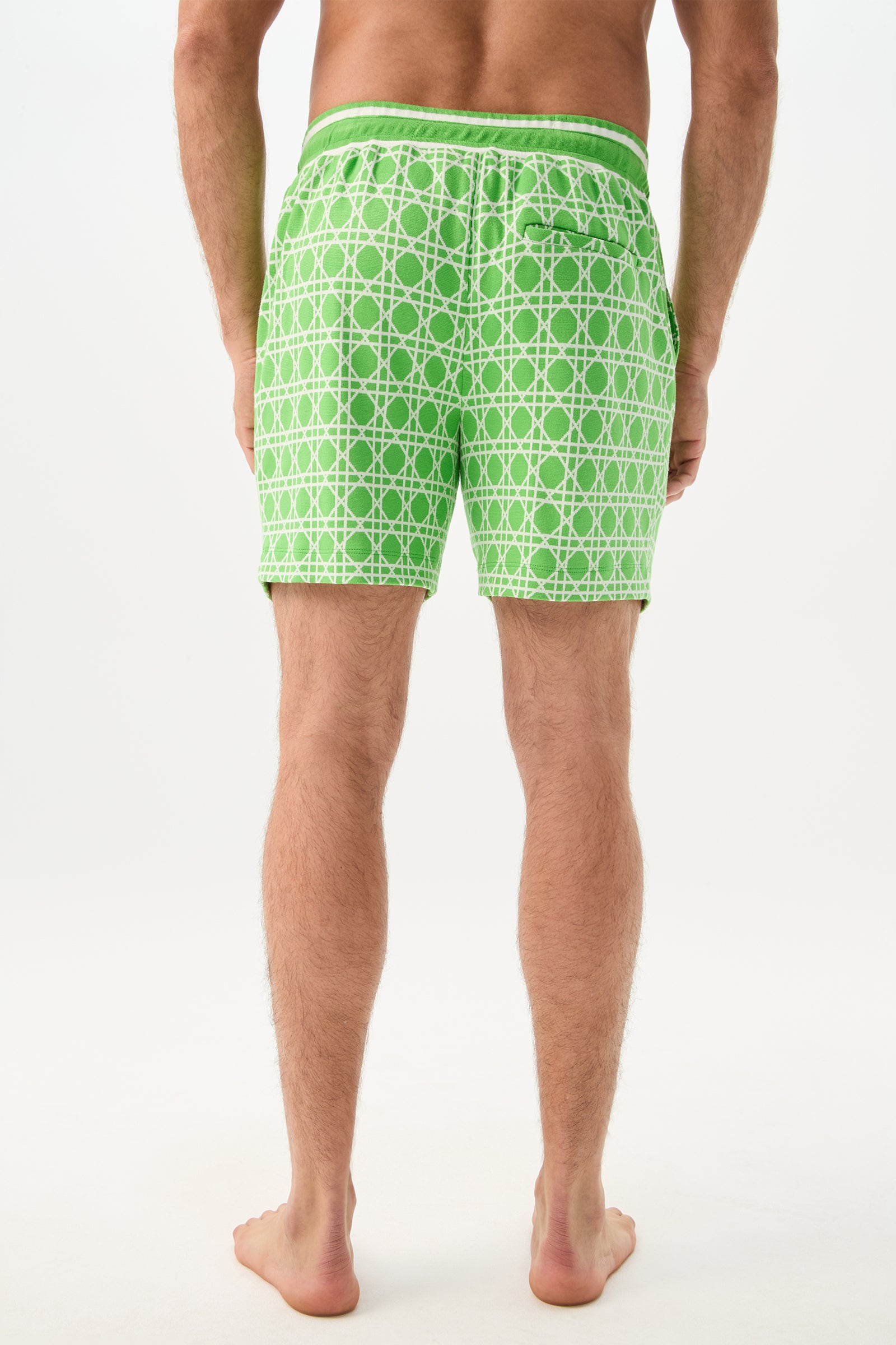 A man stands barefoot with his back to the camera, wearing the Lounge Short - Billie Jean, which features a green and white geometric pattern. The background is plain white.