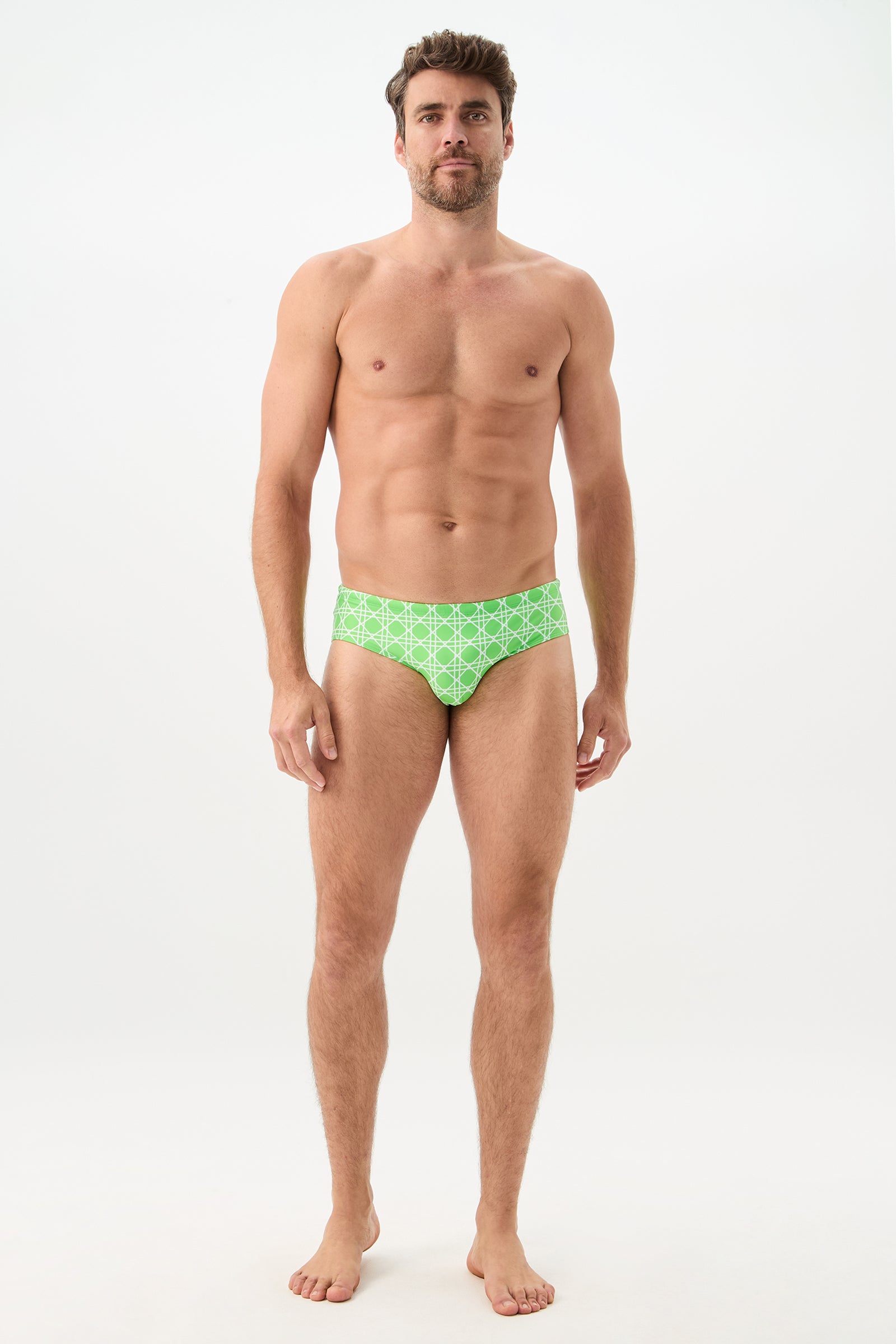 A man stands barefoot against a plain white background wearing the Bondi Swim Brief - Billie Jean. He faces forward with a neutral expression and relaxed posture, showcasing the green patterned swim briefs.