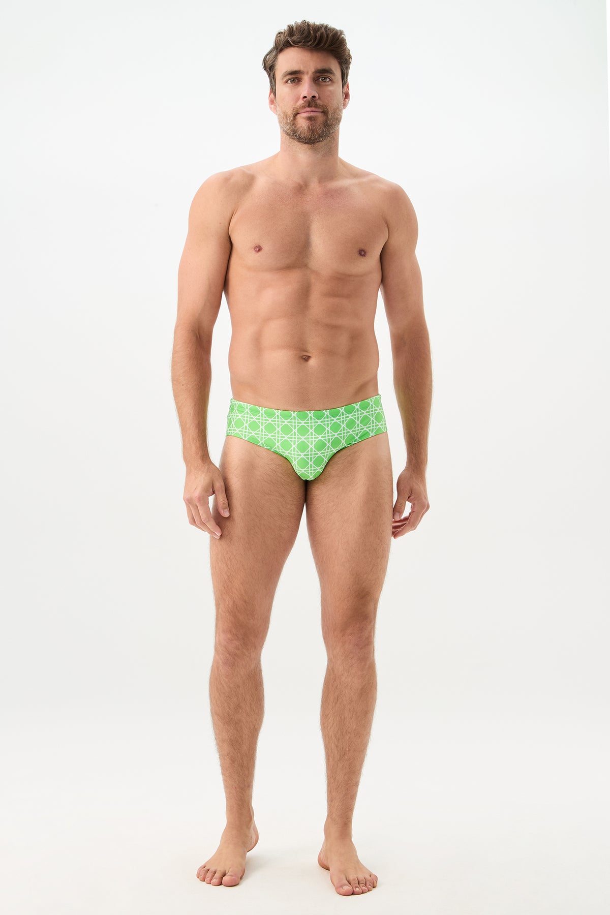 A man stands barefoot against a plain white background wearing the Bondi Swim Brief - Billie Jean. He faces forward with a neutral expression and relaxed posture, showcasing the green patterned swim briefs.
