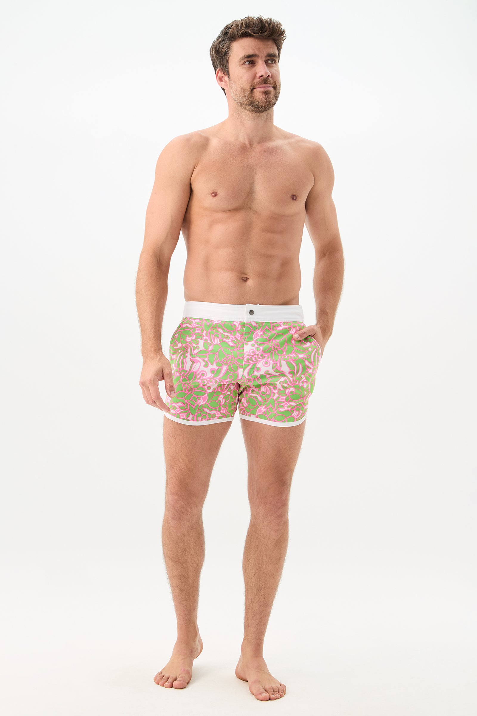 A shirtless man stands barefoot against a white background, wearing the Surfside Board Short - Petra Floral in light pink and green with white trim and a front button. He has one hand on his hip and is looking slightly to the side.