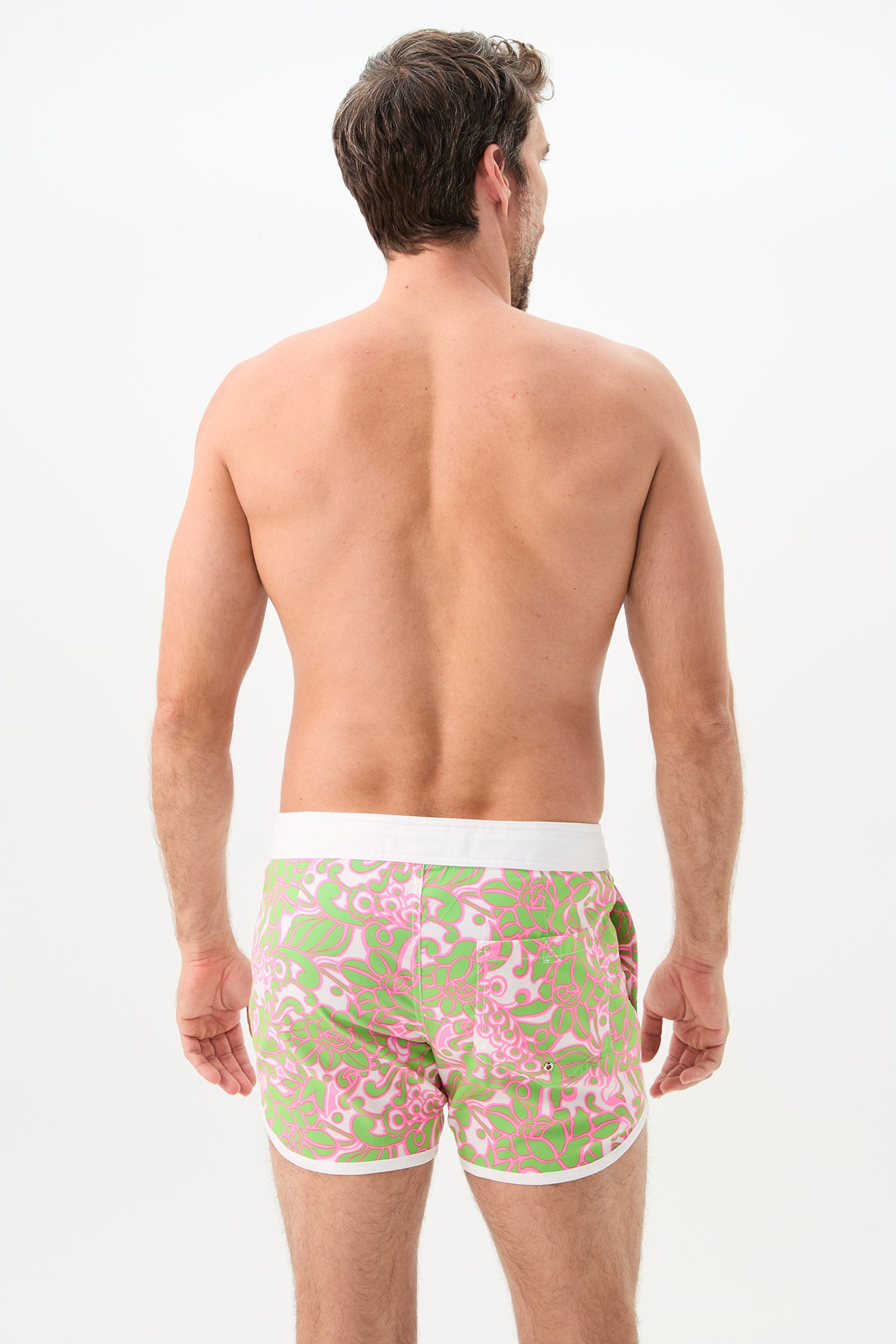 A man stands shirtless, facing away, wearing Surfside Board Short - Petra Floral with a pink and green floral pattern and white waistband, set against a plain white background.