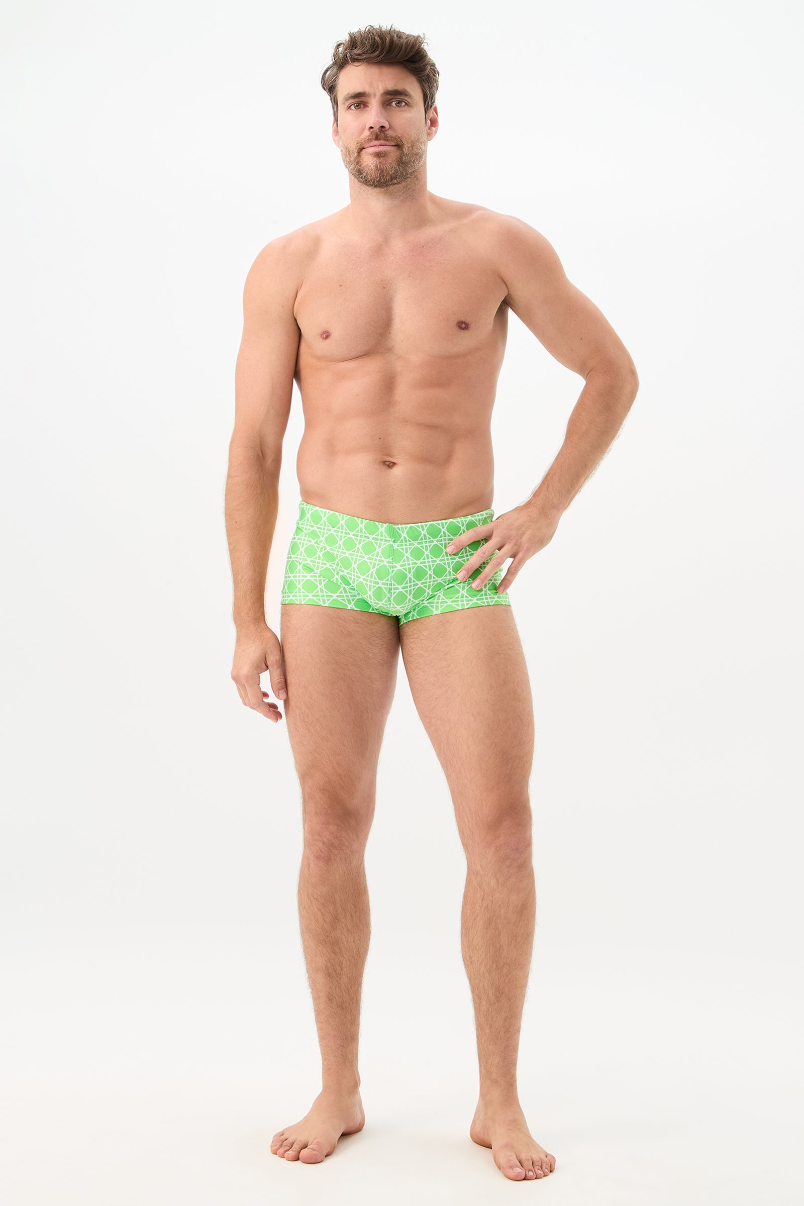 A man with short brown hair and a beard stands barefoot against a plain white background, wearing the Sorrento Swim Trunk - Billie Jean, posing with one hand on his hip and looking toward the camera.