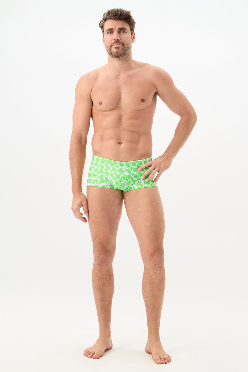 A man with short brown hair and a beard stands barefoot against a plain white background, wearing the Sorrento Swim Trunk - Billie Jean, posing with one hand on his hip and looking toward the camera.