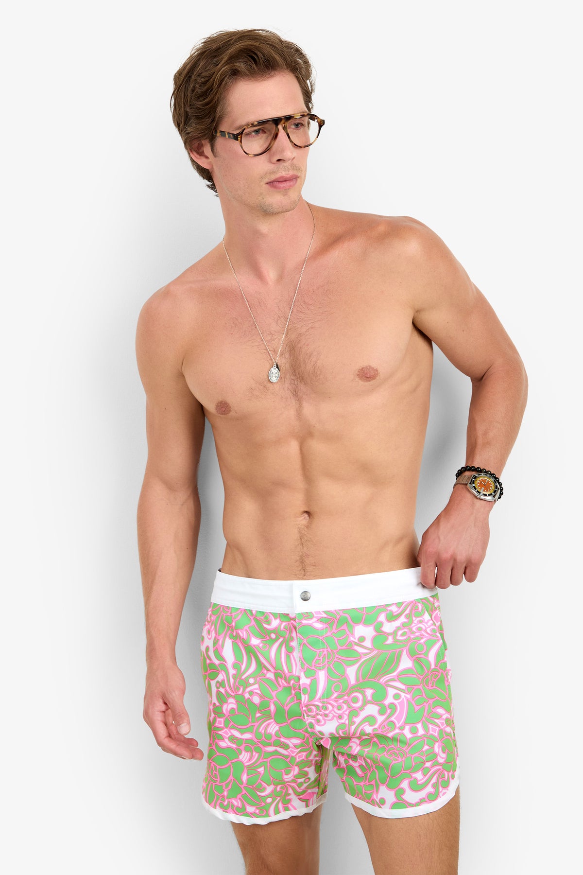 A shirtless man in glasses and a necklace wears the Surfside Boardshort, featuring pink, green, and white floral print, as he stands against a plain background looking to the side.