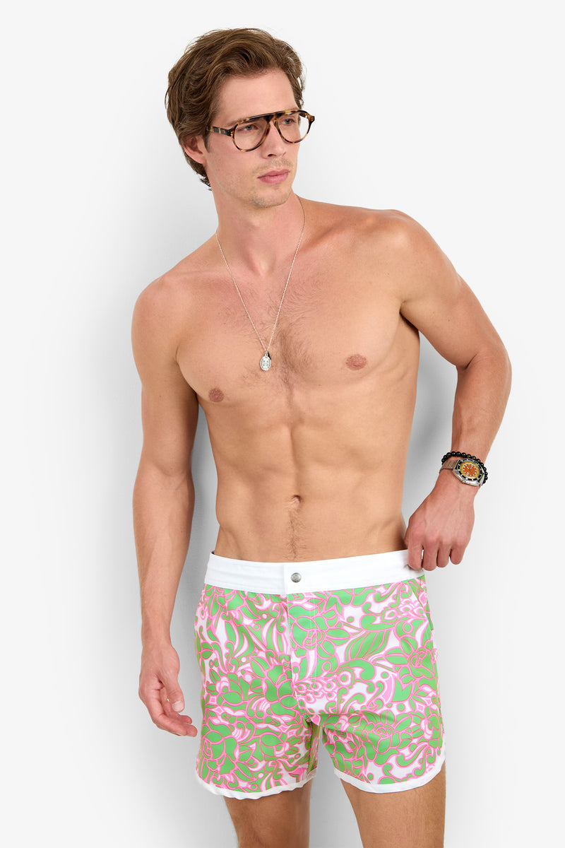 A shirtless man in glasses and a necklace wears the Surfside Boardshort, featuring pink, green, and white floral print, as he stands against a plain background looking to the side.