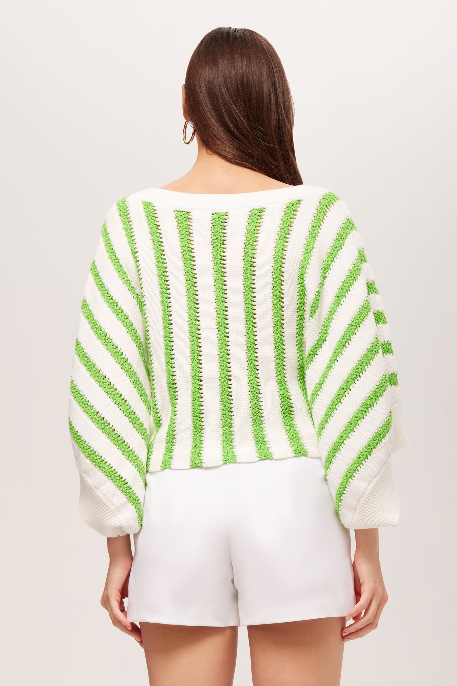 A woman with long brown hair is seen from the back wearing the Horizon 3/4-Sleeve Striped Boatneck Pullover, a white knit sweater with green stripes, paired with loose white shorts against a light background.