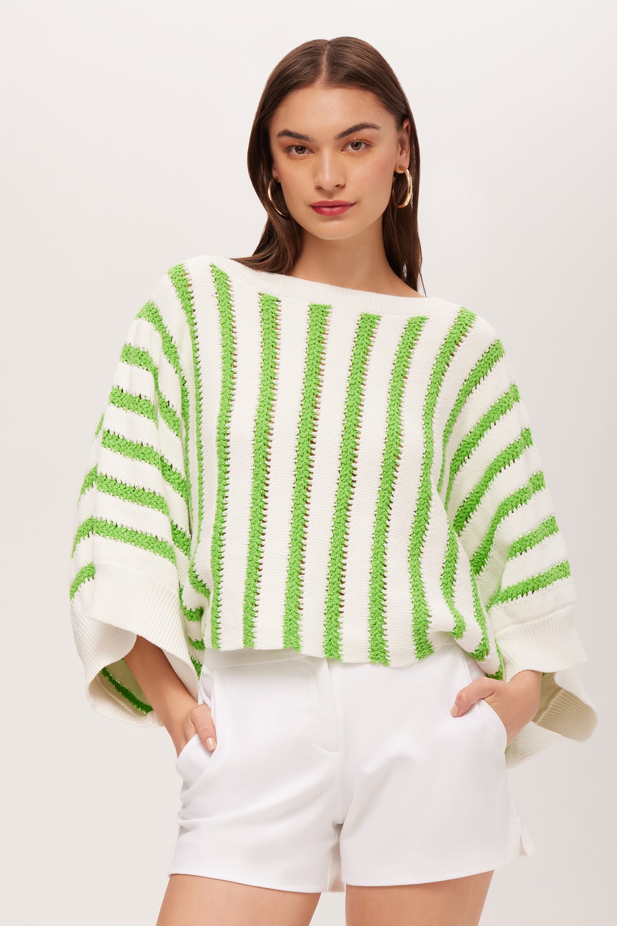 A woman stands against a plain light background, hands in pockets, wearing the Horizon 3/4-Sleeve Striped Boatneck Pullover in Serene Green/Whitewash and loose white shorts.