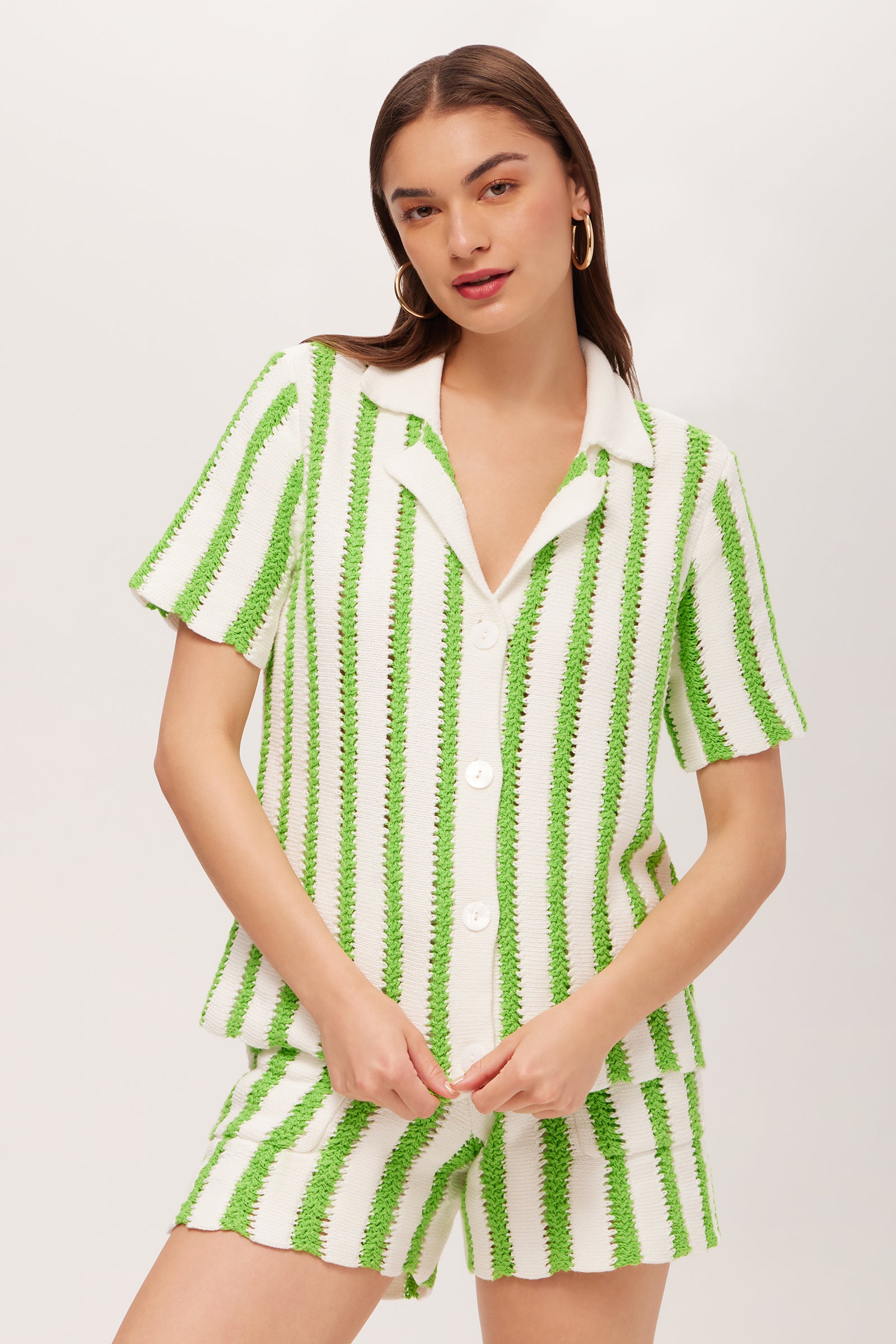 A woman poses against a plain background, wearing the Sloane Short-Sleeve Striped Button-Up Shirt in Serene Green/Whitewash, paired with matching shorts; one arm is bent while the other rests by her side.