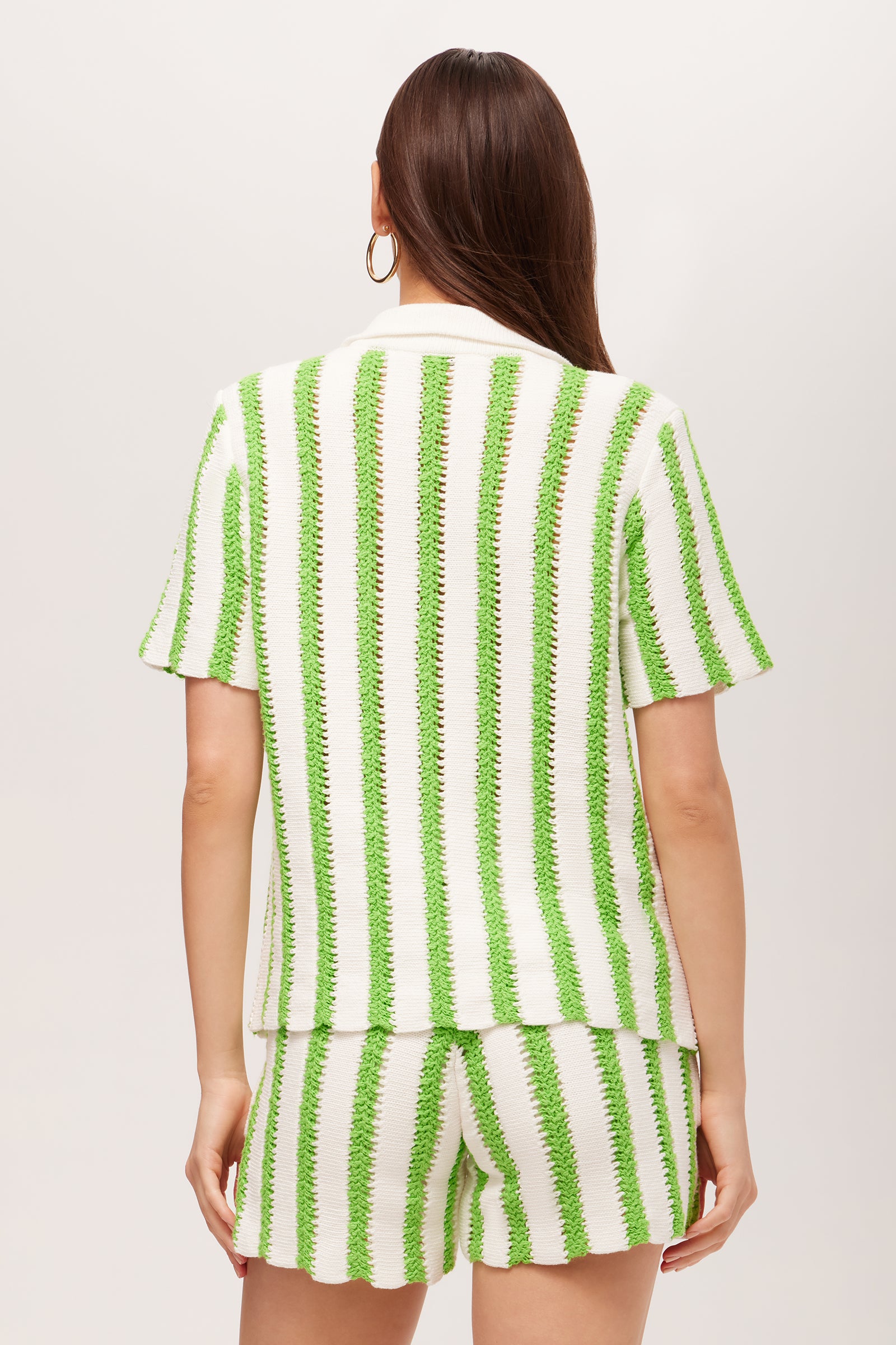 A woman with long brown hair and hoop earrings models the Sloane Short-Sleeve Striped Button-Up Shirt, a white top with vertical green stripes, shown from the back against a plain background.