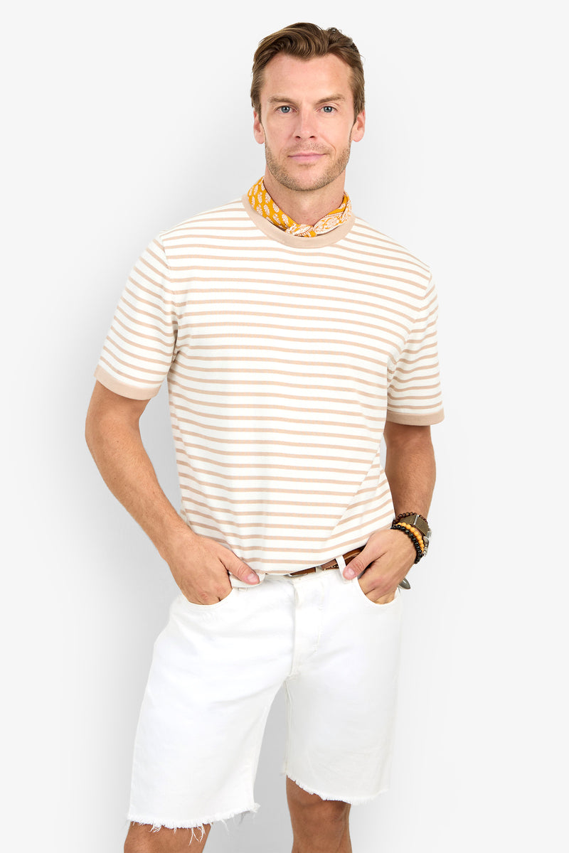 Man against a white background wears the Giovanni Stripe Short-Sleeve Pullover in beige and white stripes, paired with white frayed shorts, a yellow patterned scarf, bracelets, and smiles slightly with hands in pockets.